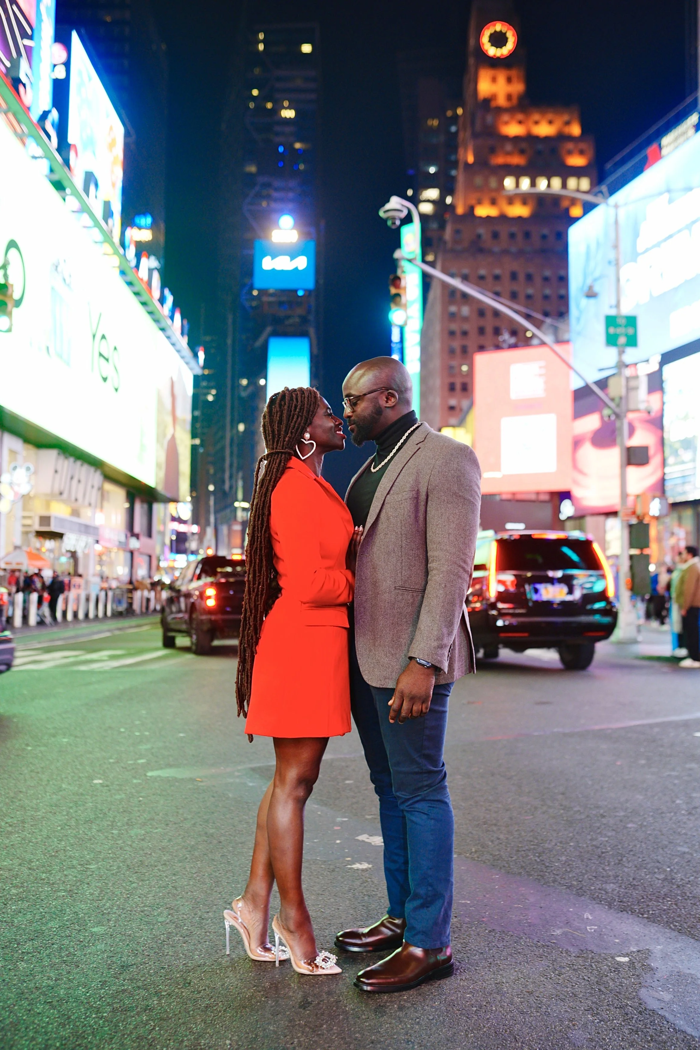 Moyo and Liam are standing close together at night in Times Square, New York City, with bright neon lights and billboards in the background. 