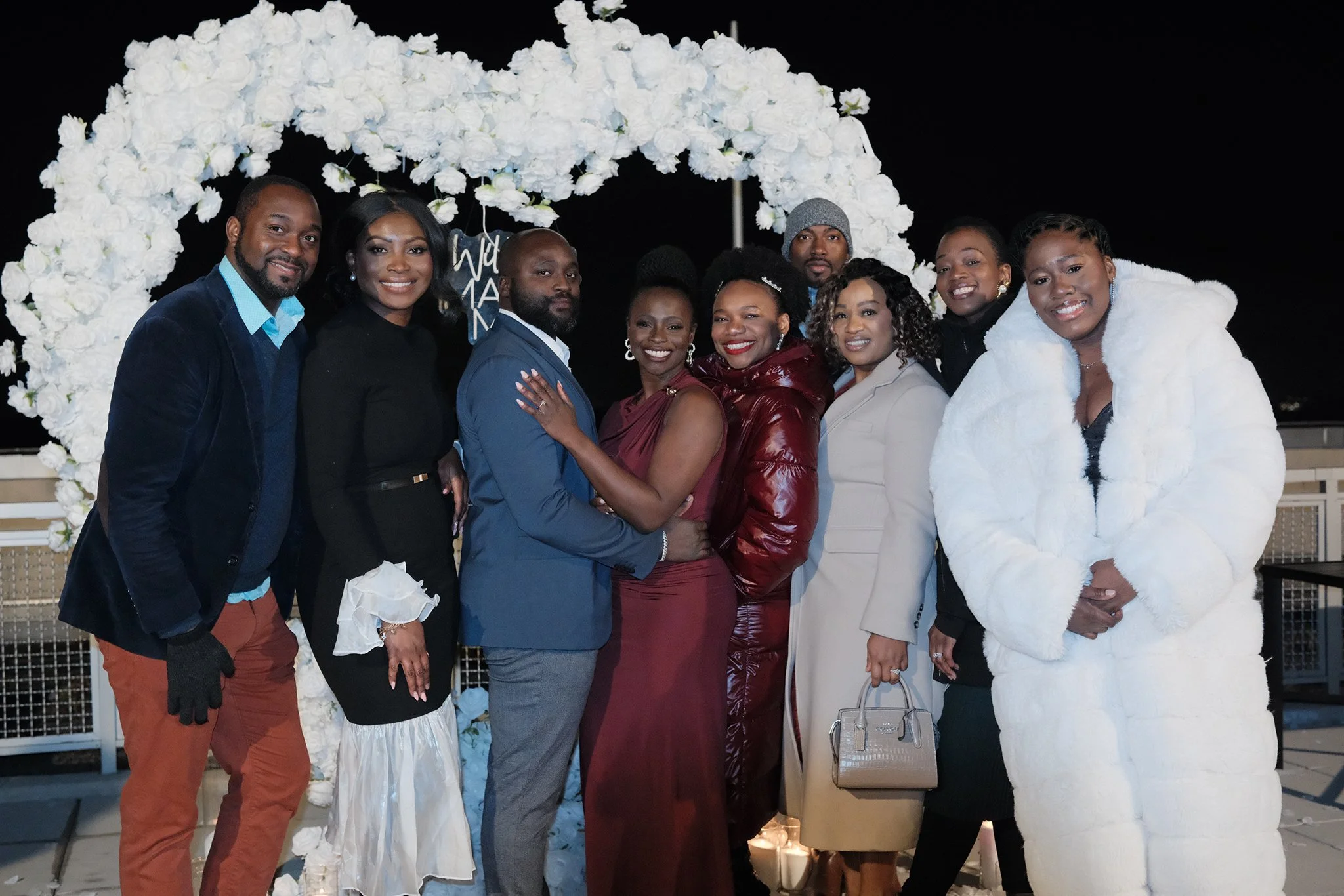 Moyo and Liam's engagement celebration, standing in front of a white floral arch under a dark night sky. They are dressed in formal and semi-formal attire, smiling and posing for the photo.