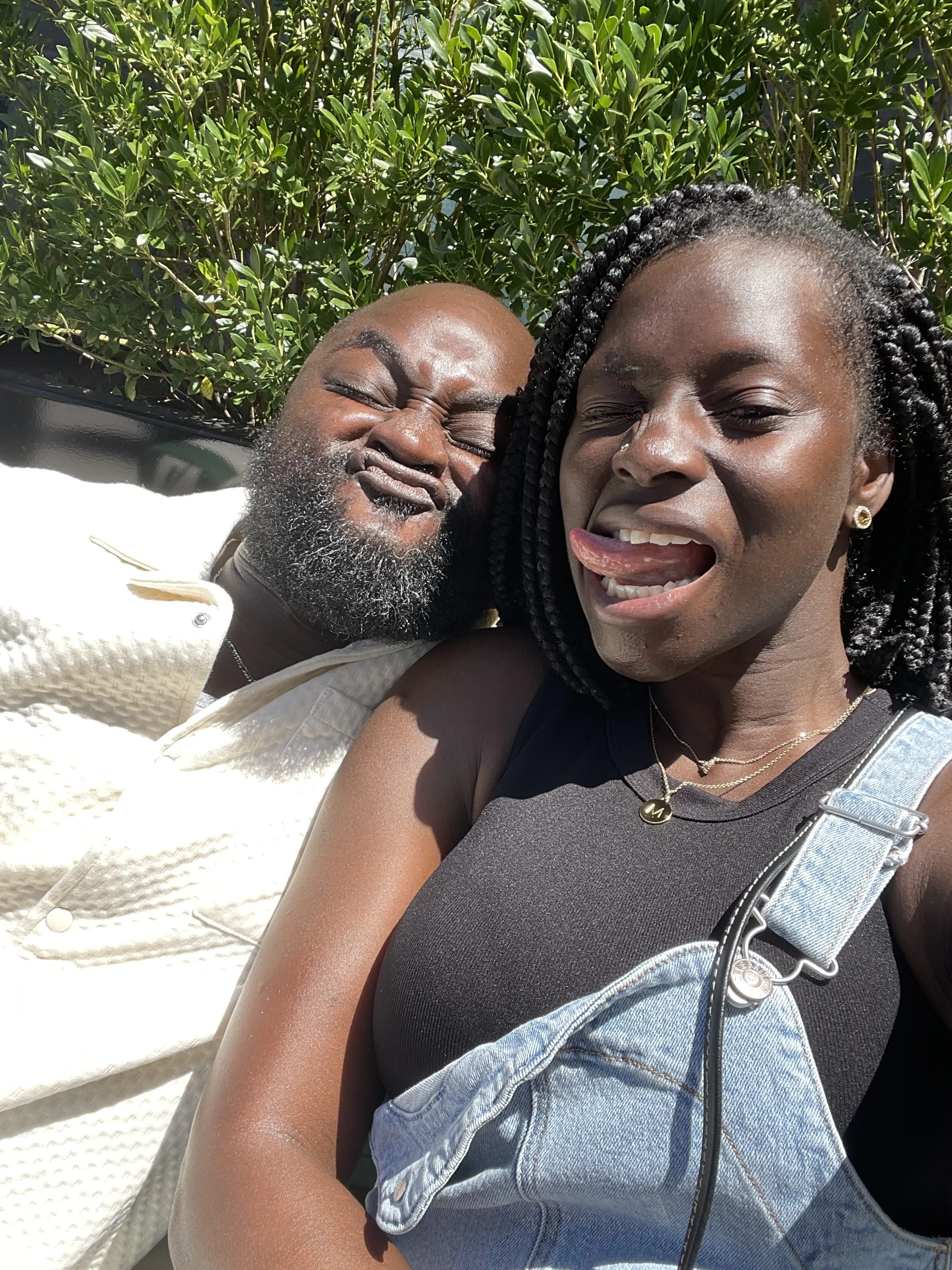 Moyo with braided hair sticking out her tongue, taking a close-up selfie with Liam with a beard and closed eyes, both outdoors with green bushes in the background.