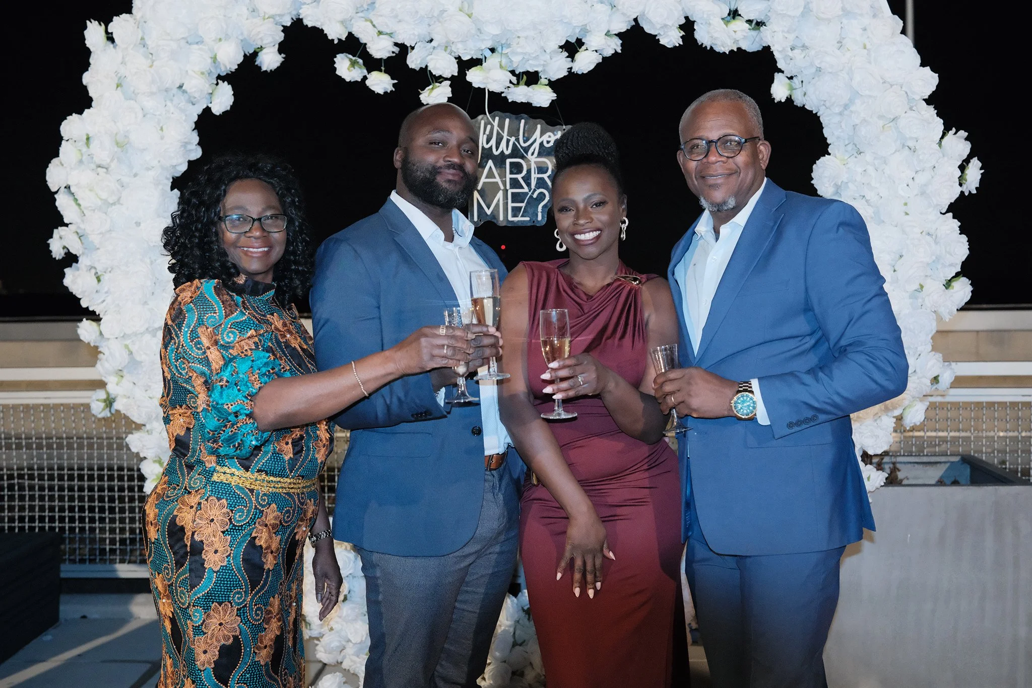 Liam and Moyo dressed formally at their engagement, standing in front of a white floral arch with a sign saying 'Will you marry me?' in the background, celebrating and holding glasses of champagne with Moyo's dad and mom.