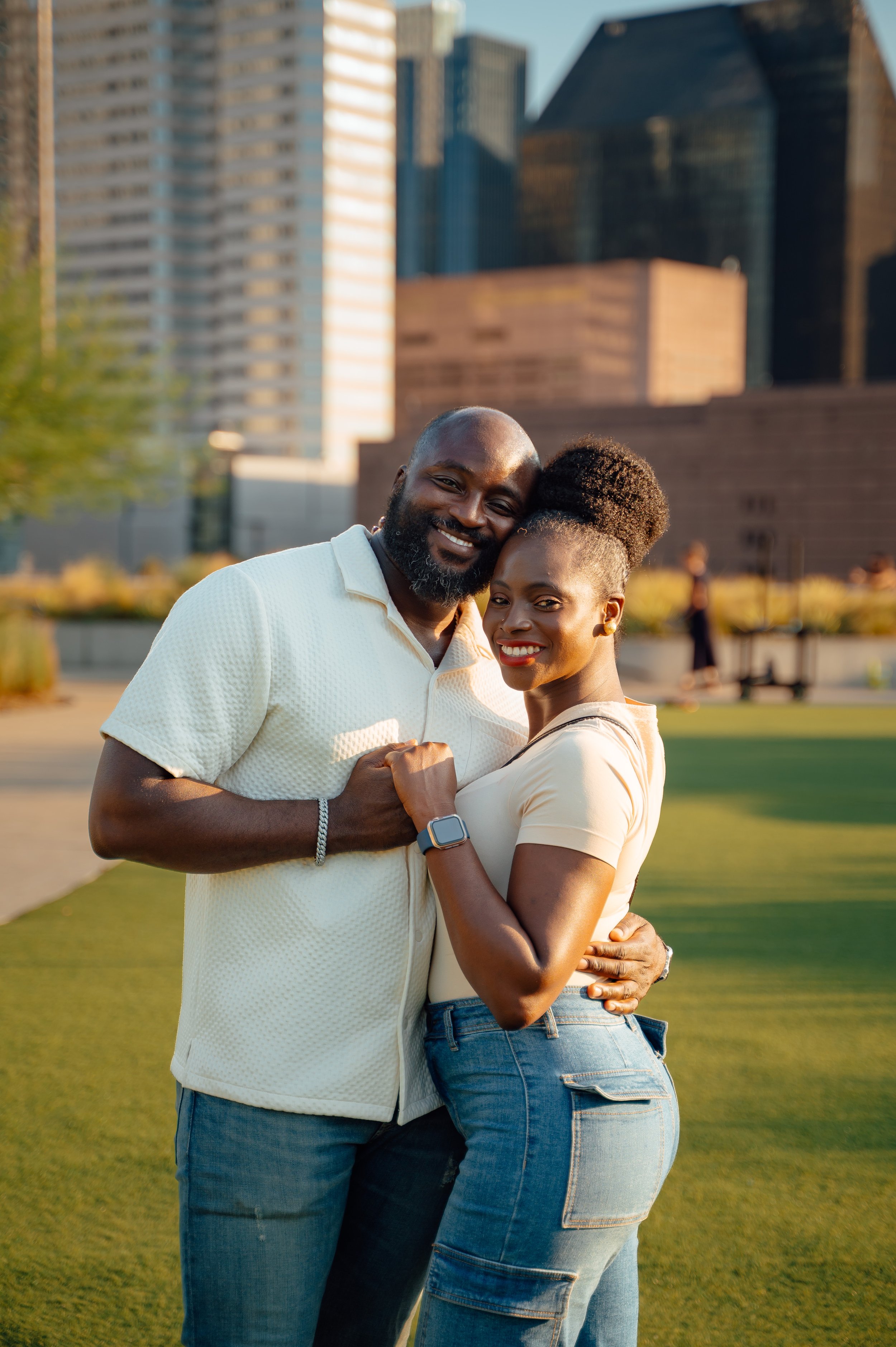 Moyo and Liam hugging outdoors in a city park with city buildings in the background during golden hour.