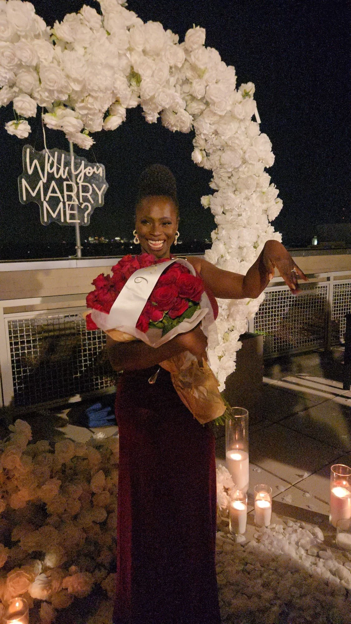 Moyo is standing outdoors at night, smiling and holding a bouquet of red roses wrapped in paper. Behind her, there is a decorative arch made of white flowers and a sign that says 'Will you MARRY ME?'