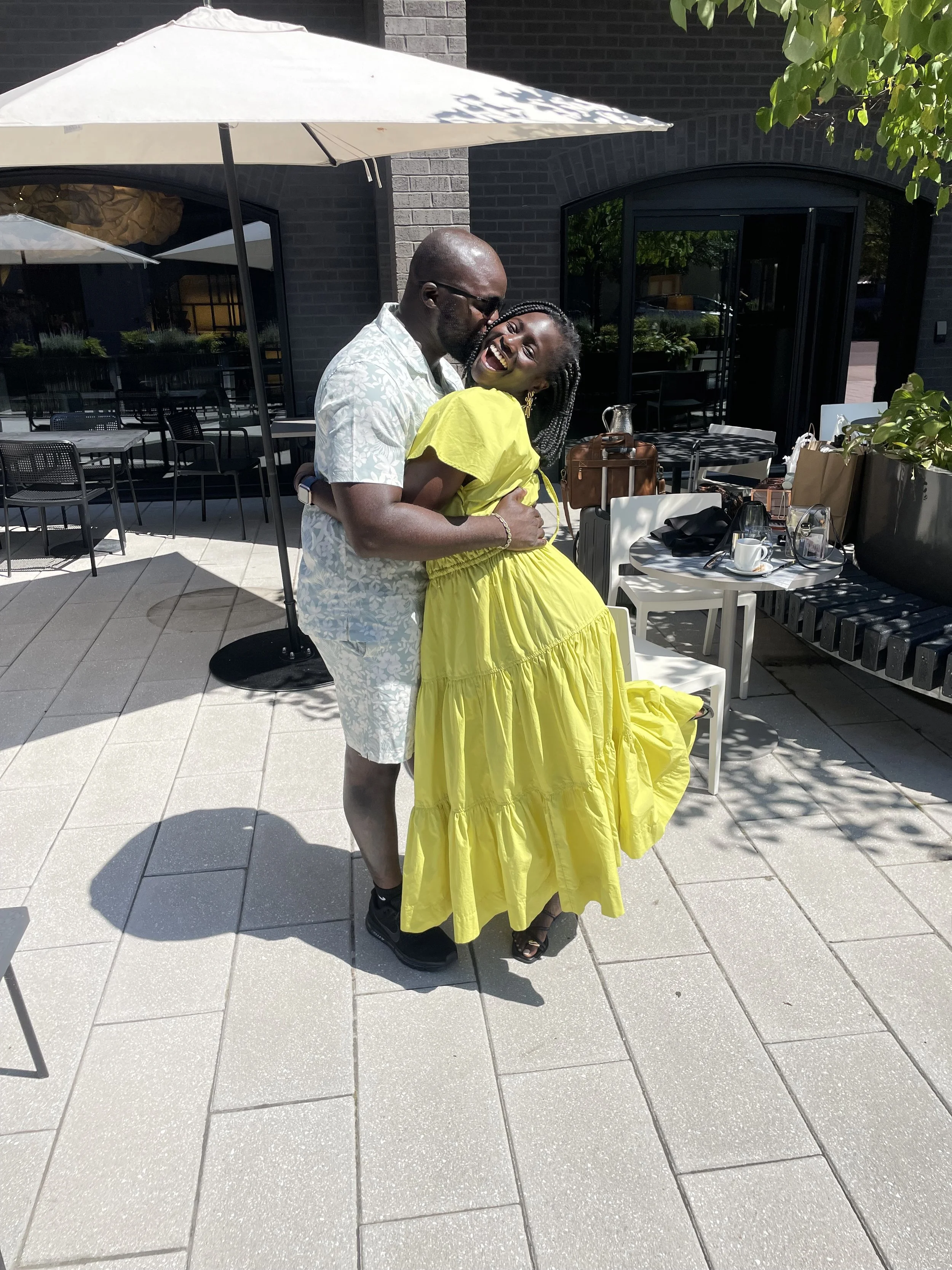 Liam with a patterned T-shirt and sunglasses, and Moyo with a bright yellow dress, smiling and hugging outdoors under a large white umbrella on a patio with tables and chairs. 