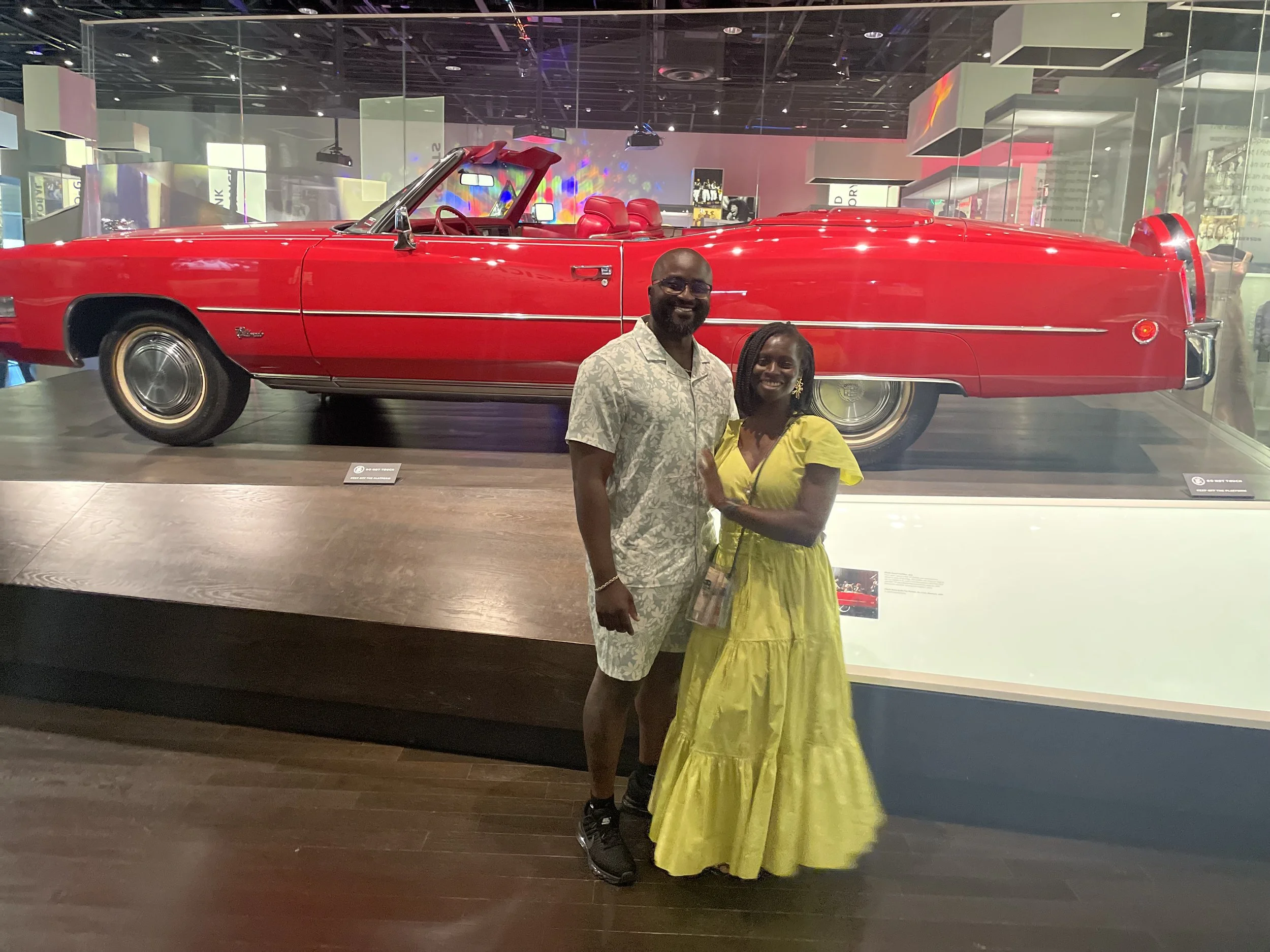 Moyo and Liam standing in front of a red vintage convertible car in a museum or exhibition space.
