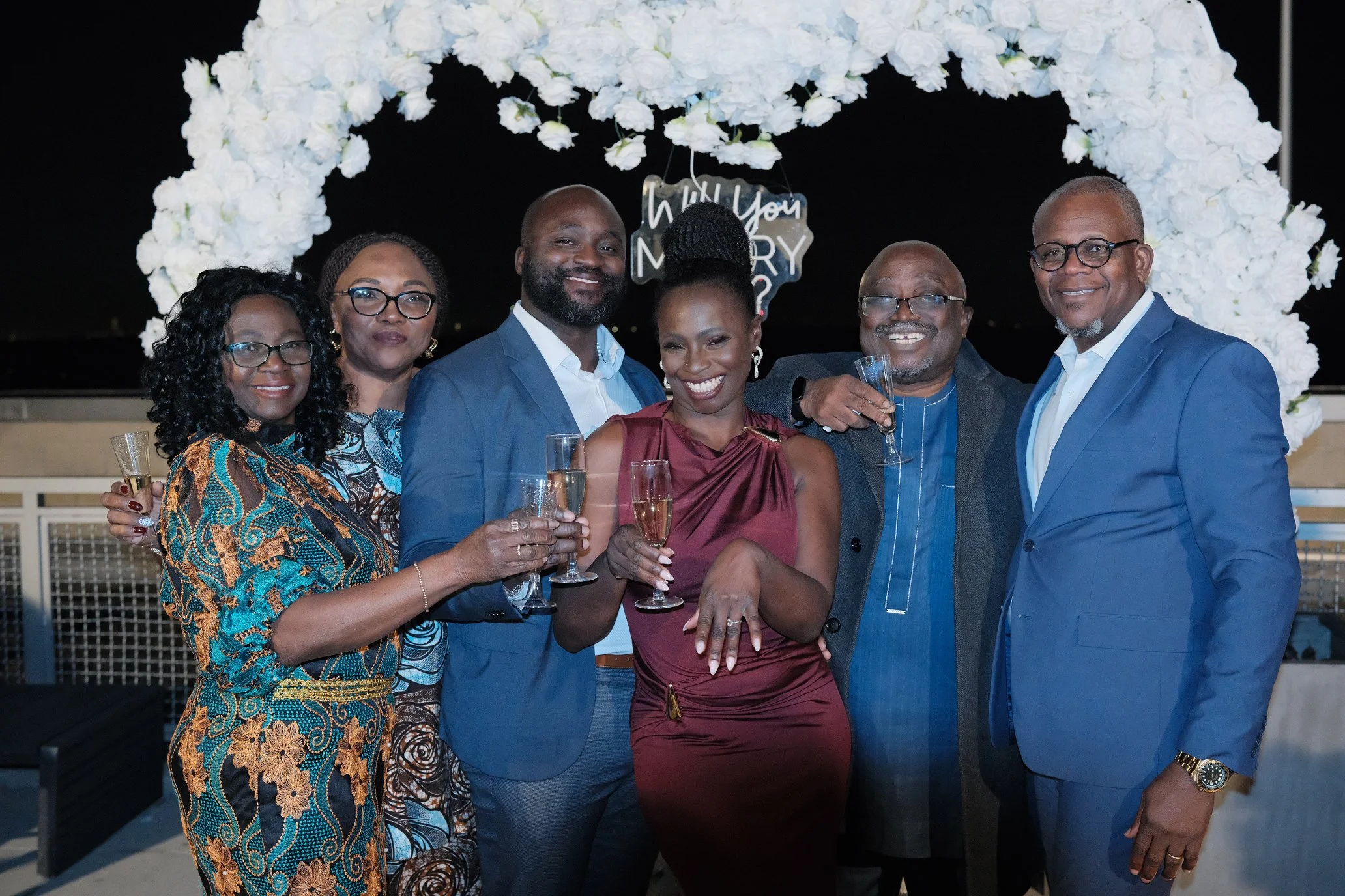 Liam and Moyo, dressed in formal attire, stood together at their engagement celebration, smiling with their parents.