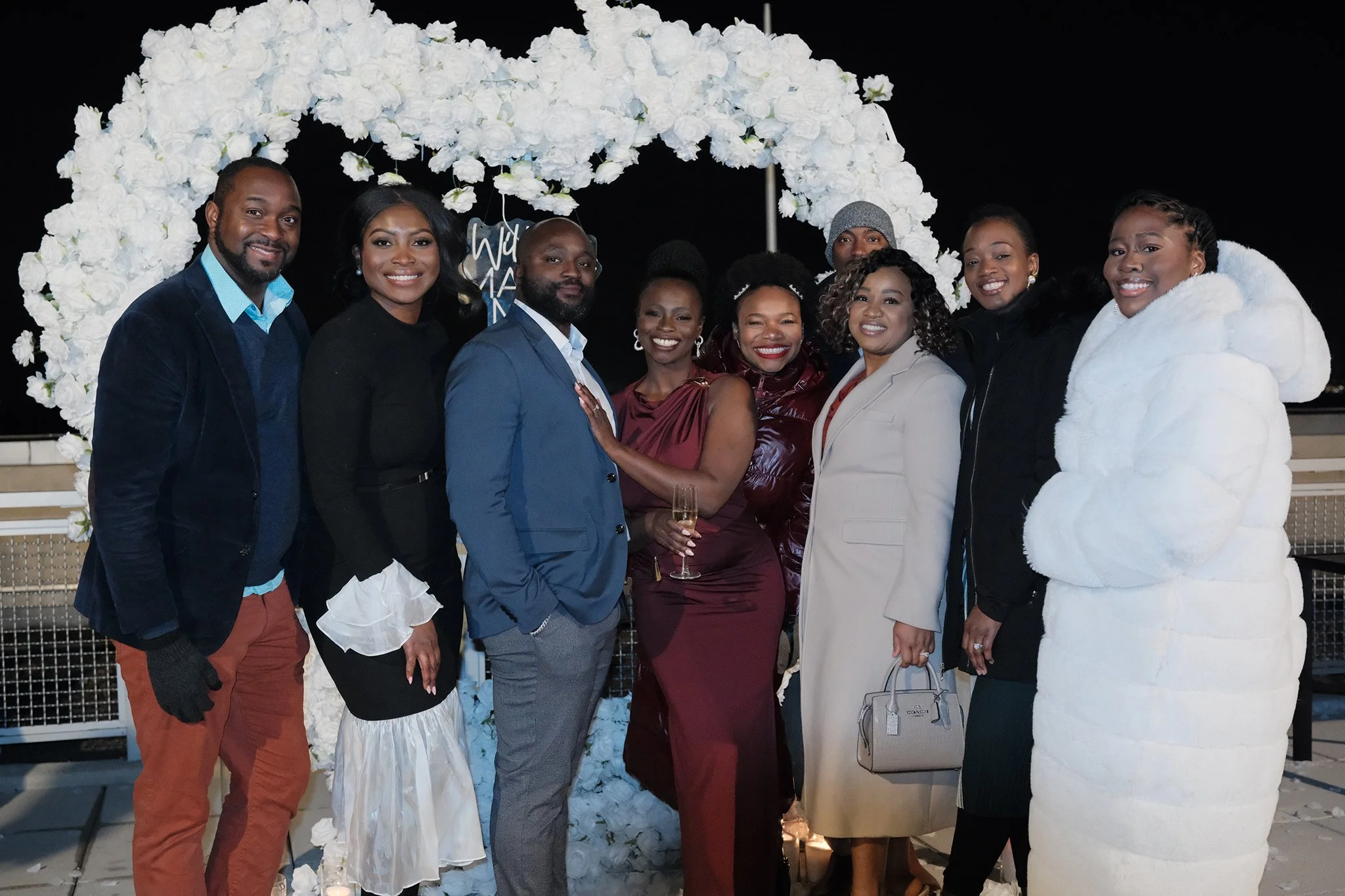 Moyo and Liam's engagement celebration at night under an arch of white roses, with some holding drinks, smiling, and dressed in formal and casual attire, with their friends.