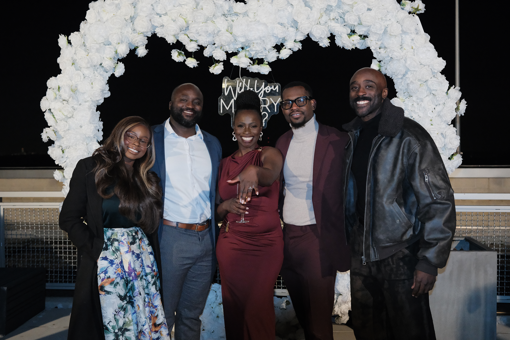 Moyo and Liam at their engagement celebration, standing in front of a heart-shaped floral arch with white roses. Posing with their siblings.
