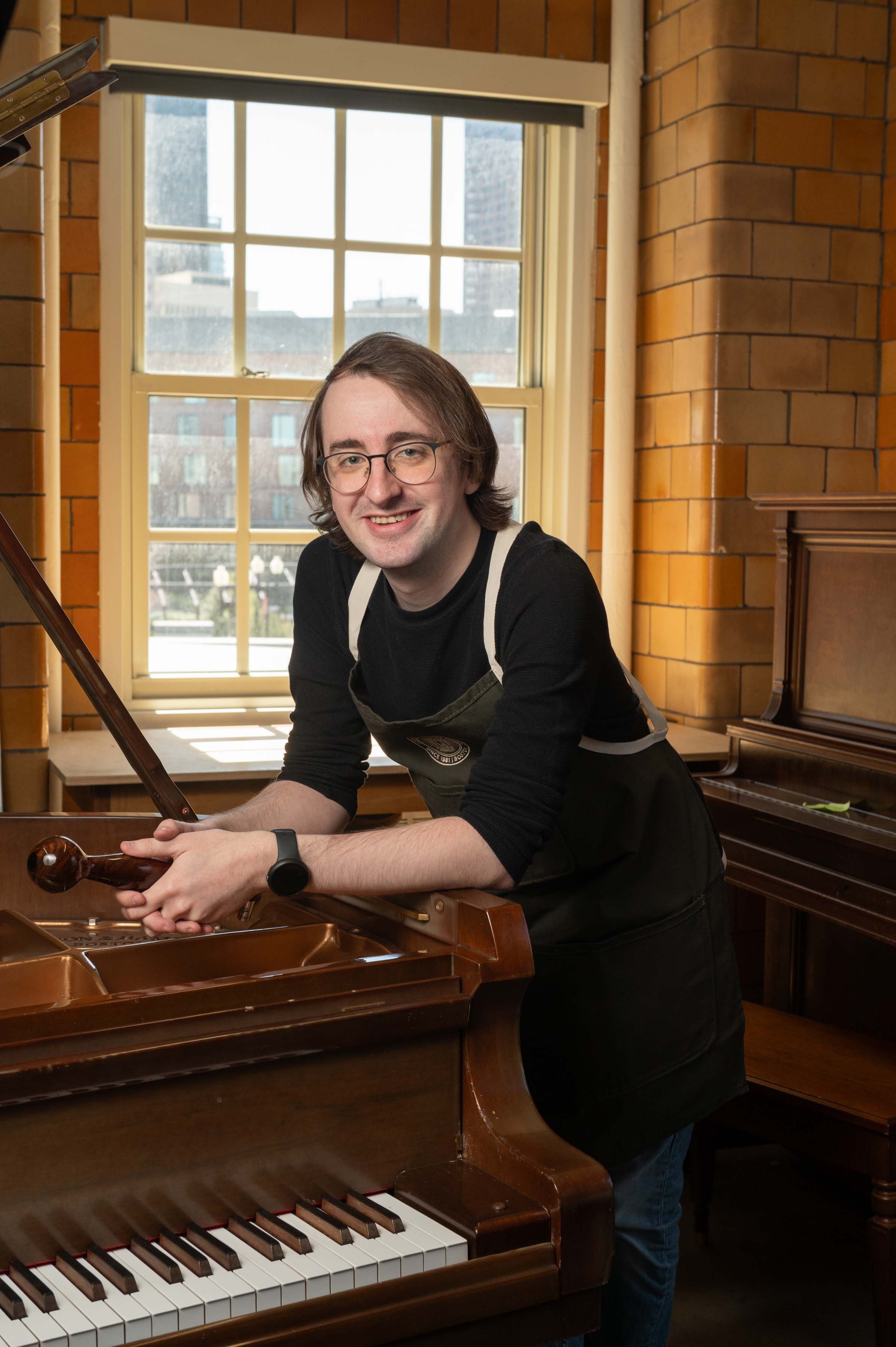 Orlin Twig, A Young man with long brown hair leaning and posing against a piano, smiling.