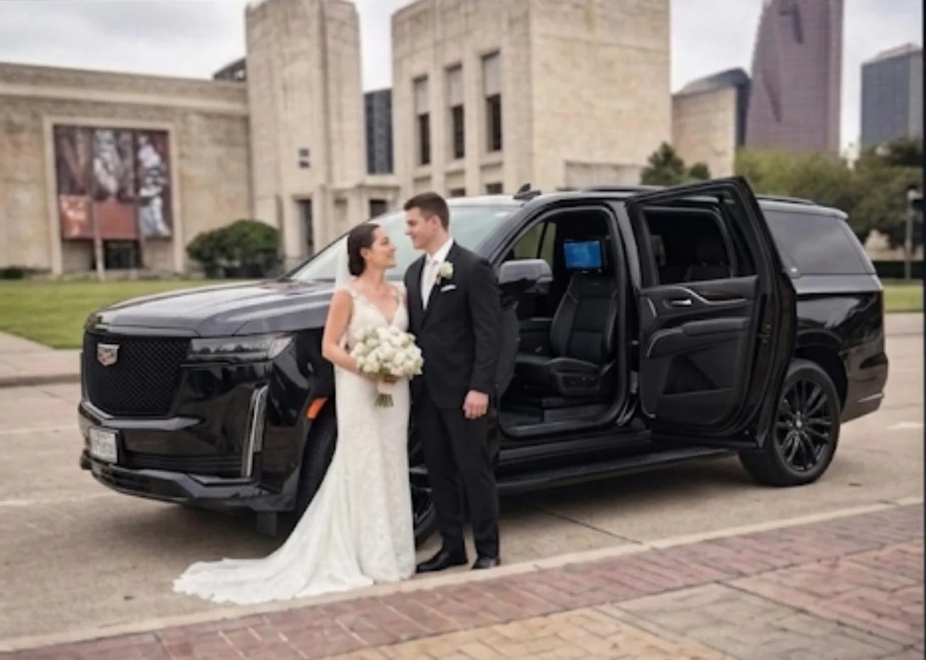 A bride and groom standing in front of a black Cadillac limousine on a city street with a large government or historic building in the background. The bride is in a white wedding gown holding a bouquet of white roses, and the groom is in a black tuxedo.