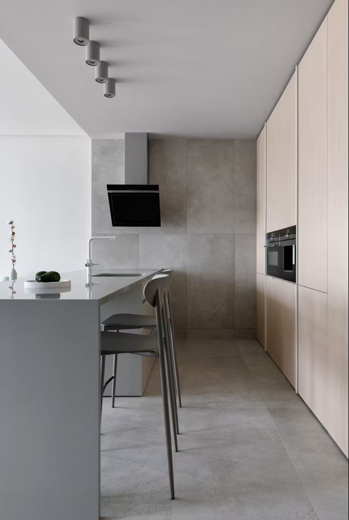 Minimalist kitchen with light wooden cabinets, built-in oven, gray tiled walls, and a gray countertop with two chairs, a small vase with flowers, and a black range hood.
