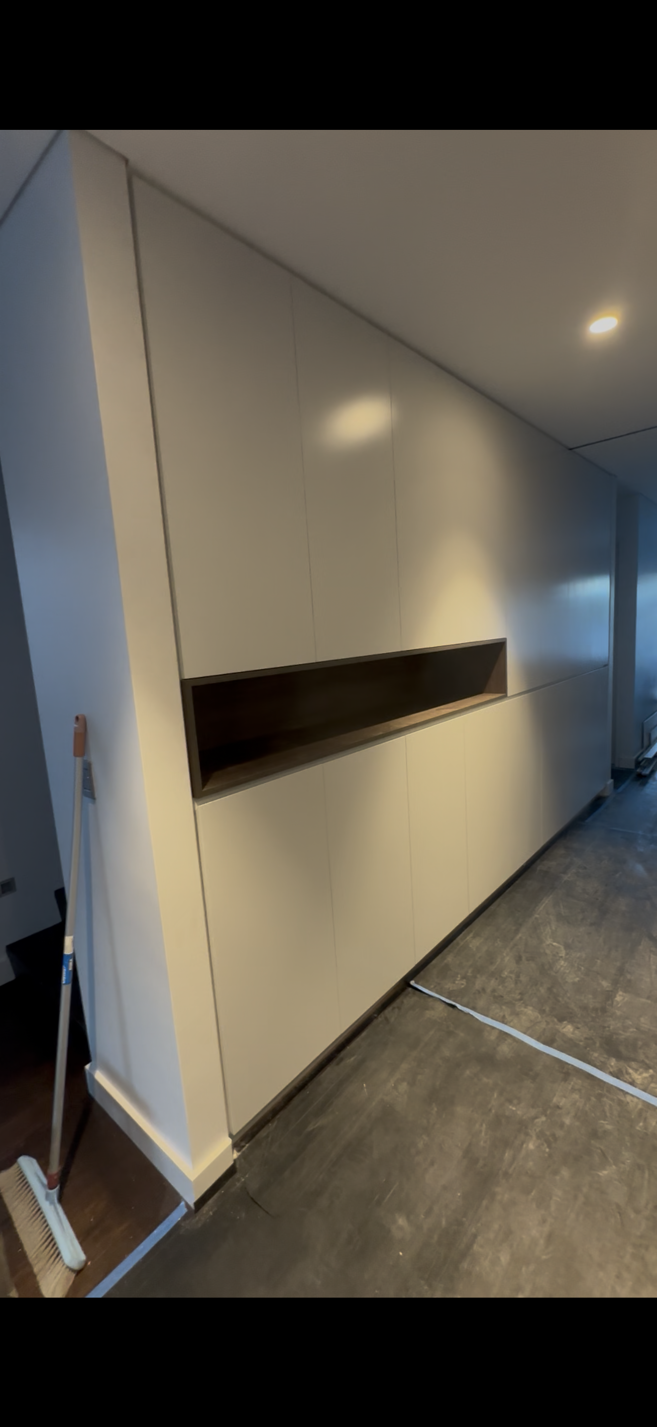 Interior view of a modern, minimalist built-in cabinet with a dark open shelf, situated against a beige wall with ceiling spotlights.