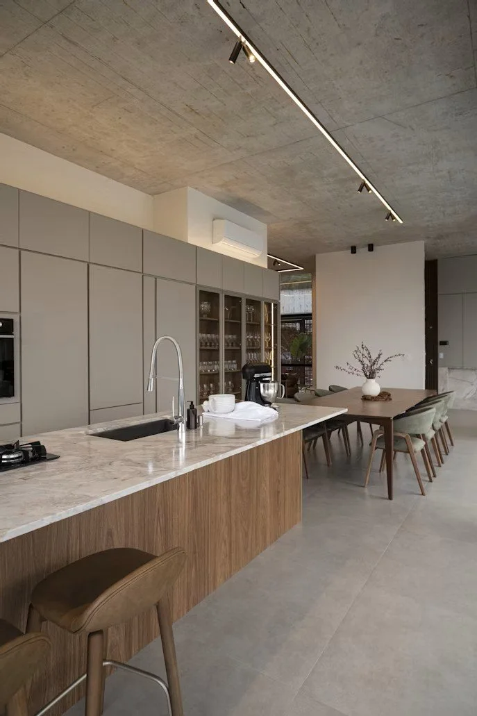 Modern kitchen with marble island, gray cabinets, and a dining area with a wooden table and green chairs, decorated with a vase of flowers.