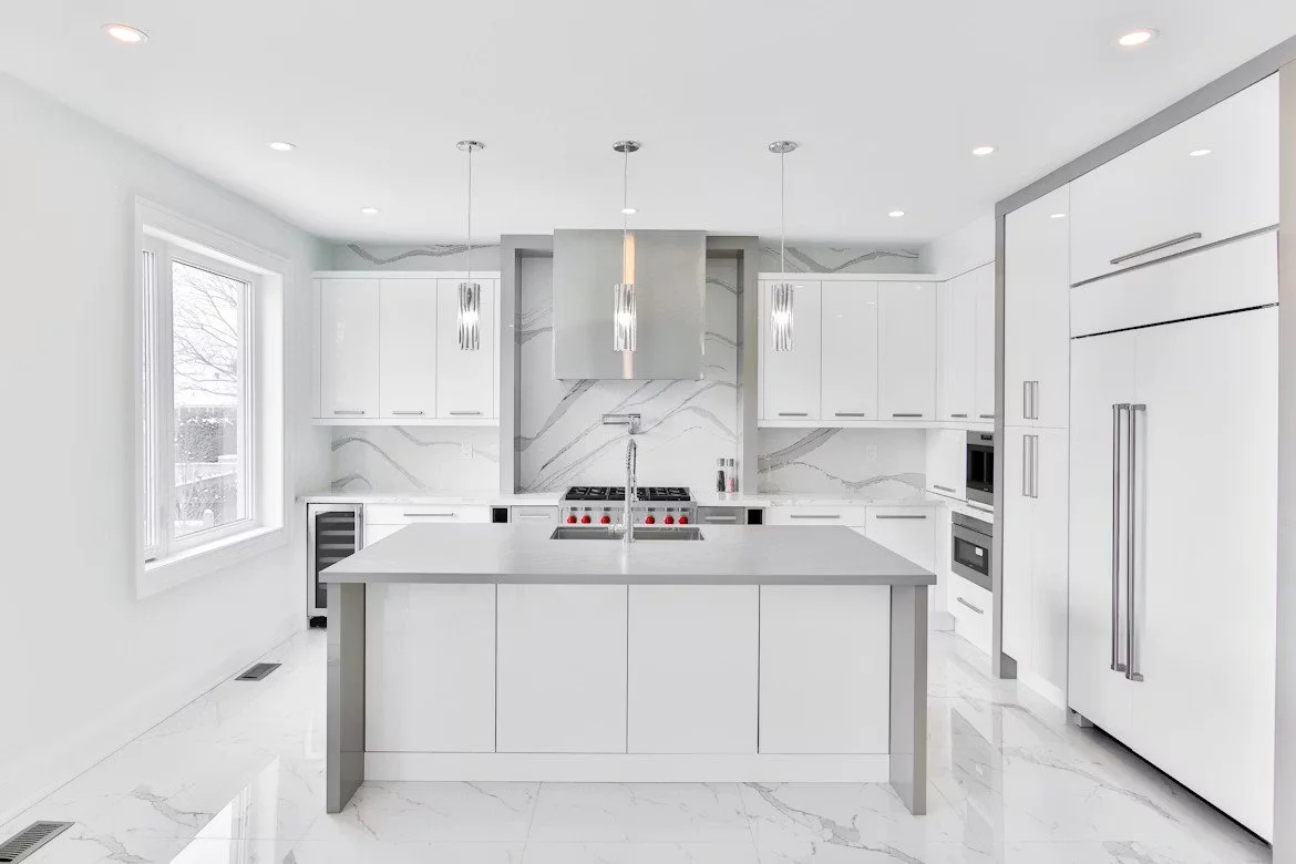 Modern white kitchen with marble countertops, an island with a sink, stainless steel range hood, and sleek appliances including a built-in microwave and refrigerator. Pendant lights hang above the island.