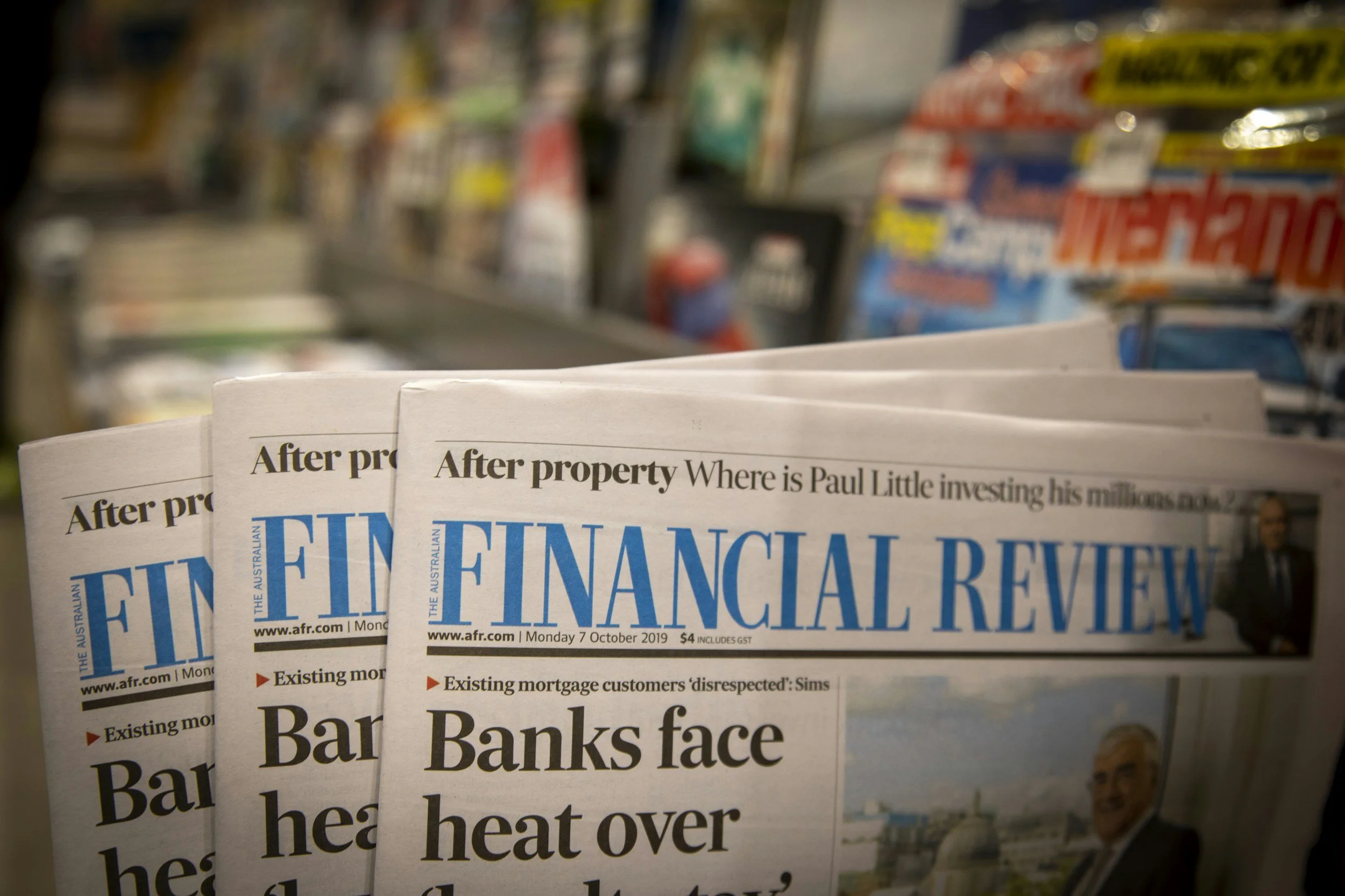 Close-up of multiple folded newspapers titled 'Financial Review' with headlines about banks facing heat over property issues, on a store shelf with a blurred background of miscellaneous products.