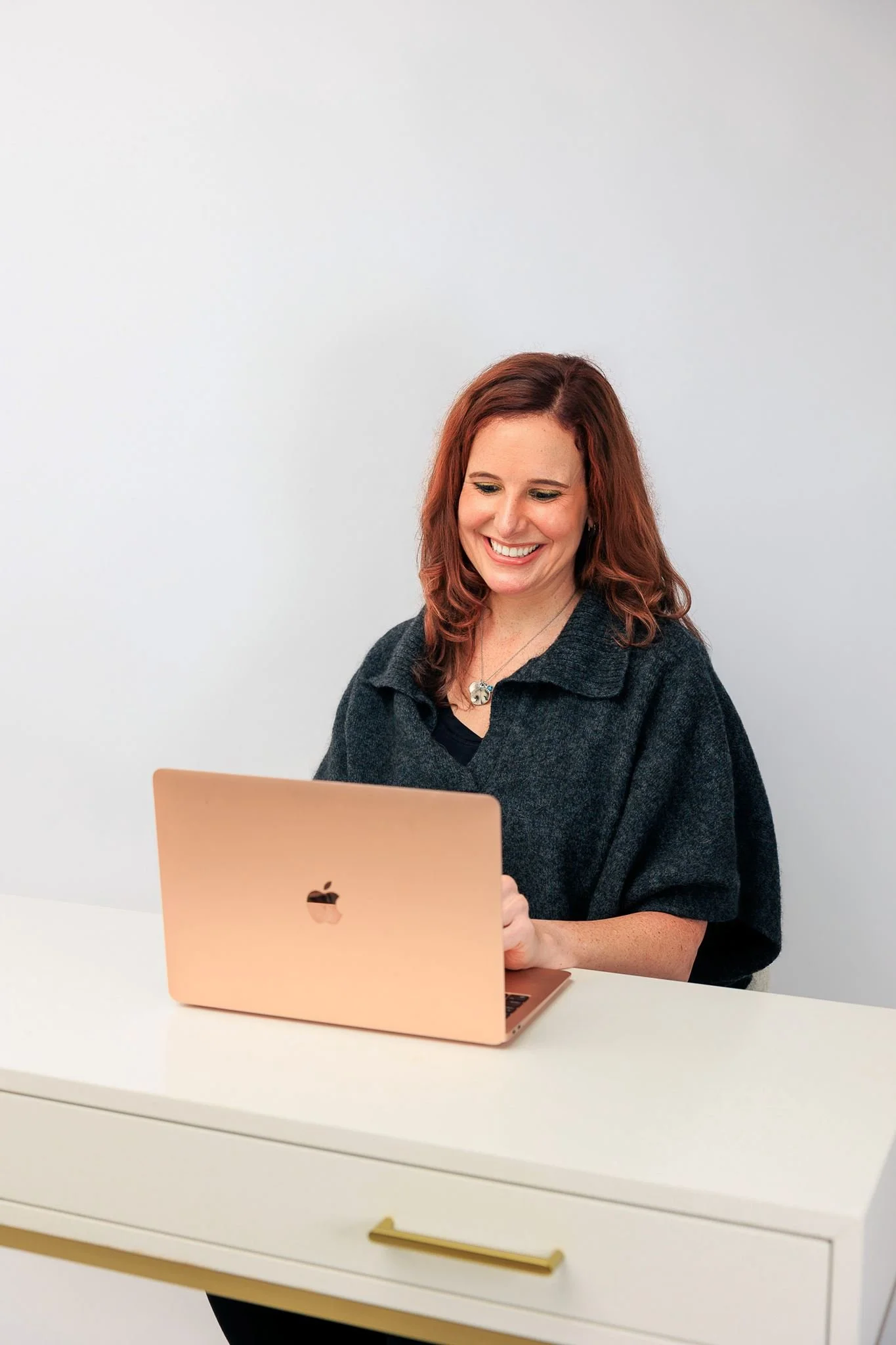 A woman with reddish-brown hair sitting at a white desk, smiling while looking at a rose gold Apple MacBook laptop.