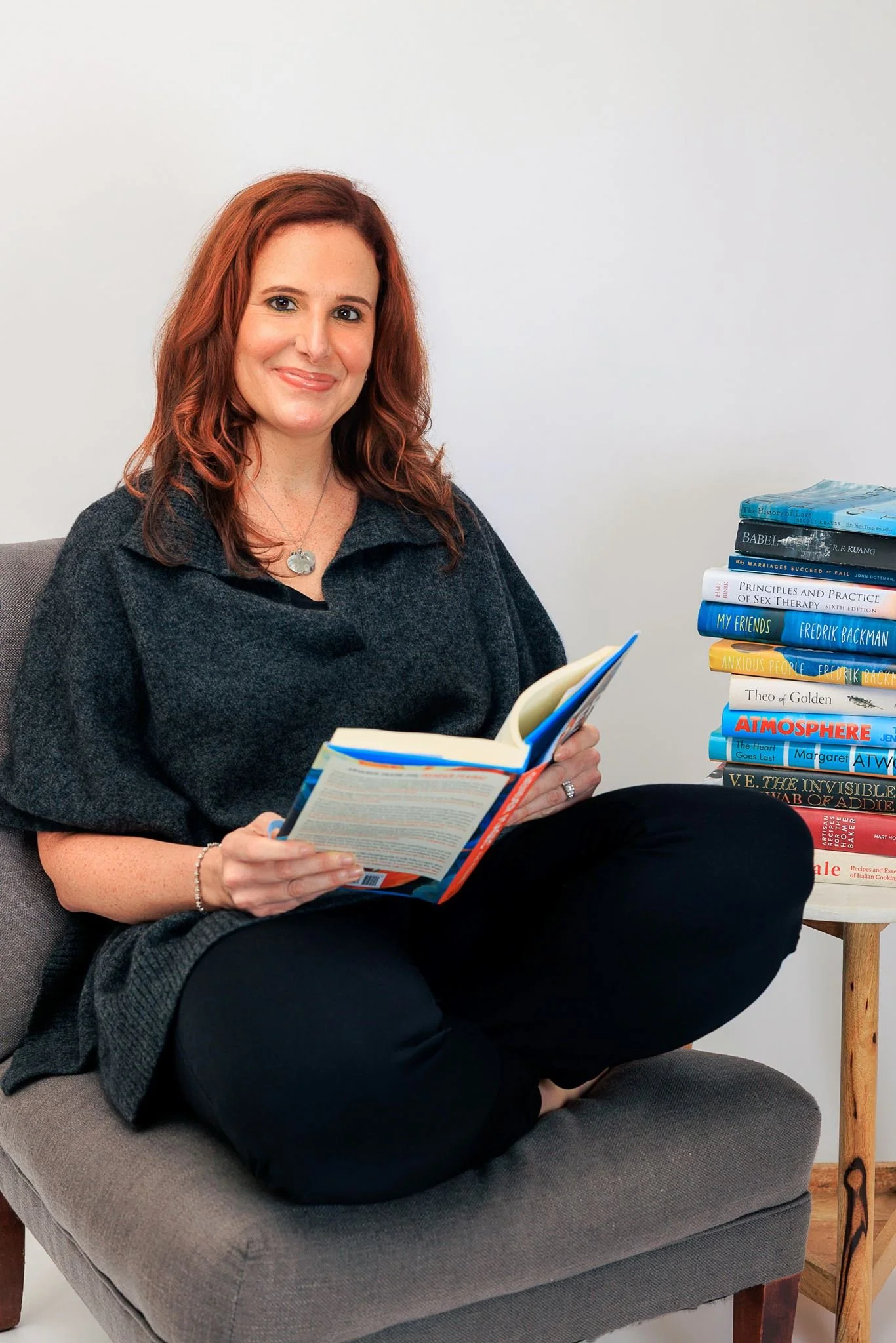 A woman with red hair sitting on a gray couch, reading a book, with a stack of books on a side table next to her.