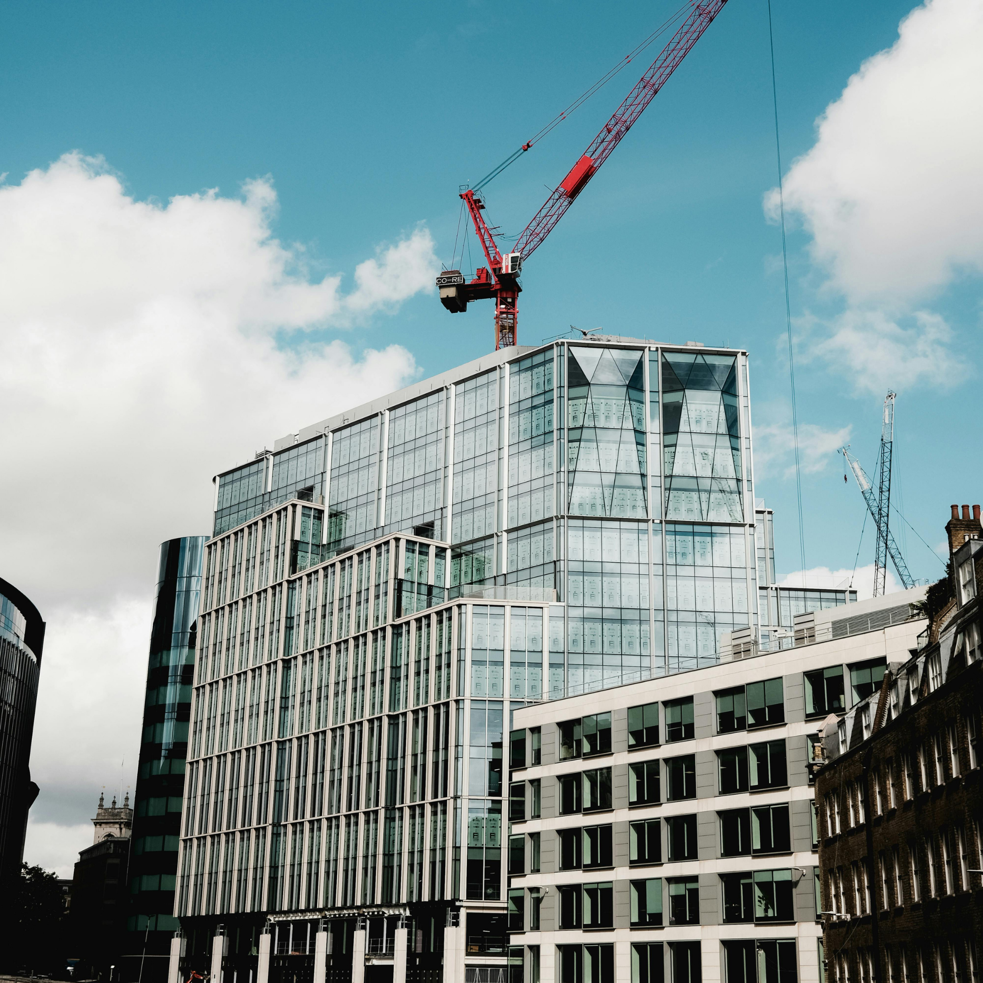 A modern glass-fronted high-rise building under construction with a red tower crane on top against a partly cloudy sky.