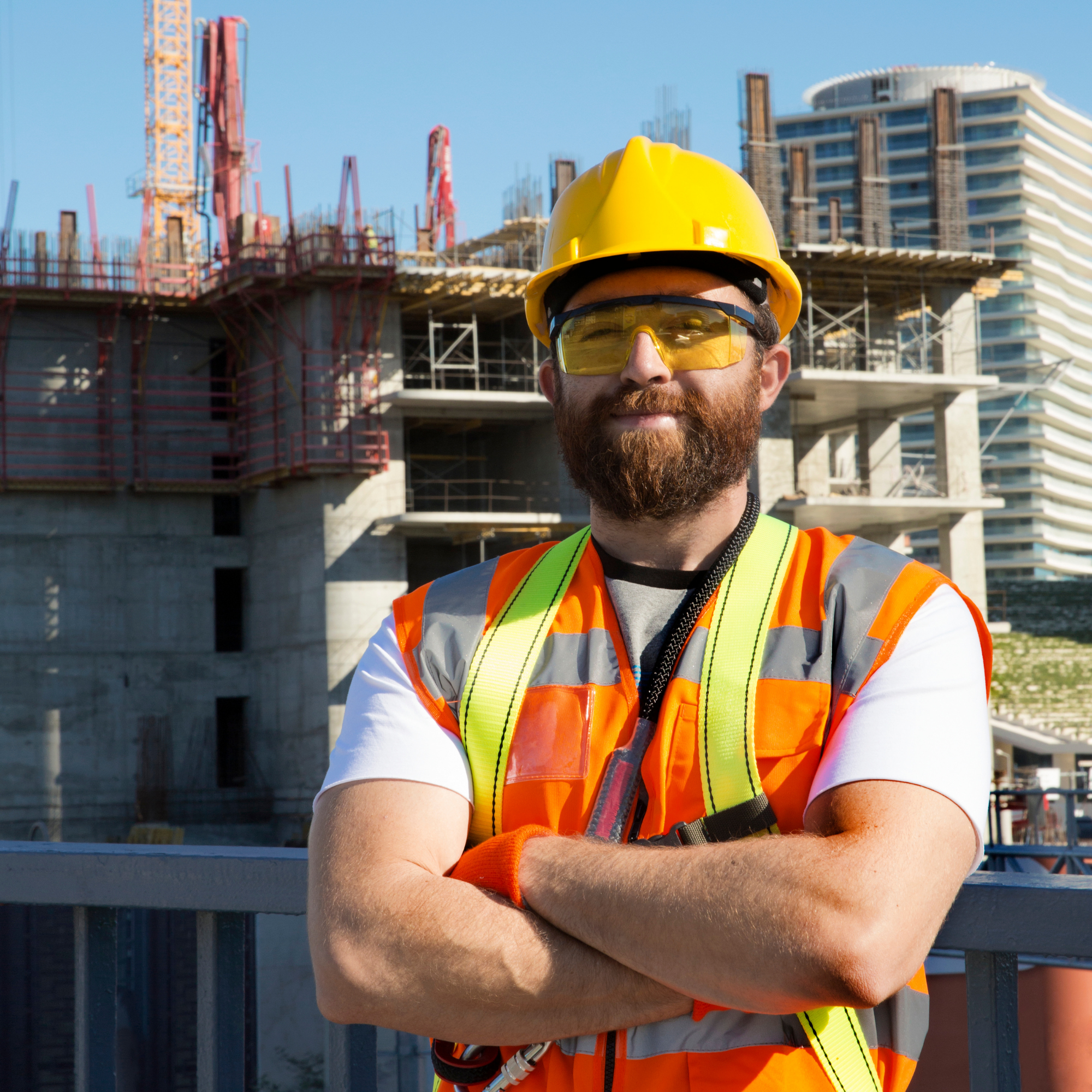 Construction worker wearing a yellow safety helmet, yellow safety glasses, a high-visibility orange safety vest, and a gray shirt standing at a construction site with building frameworks and cranes in the background.
