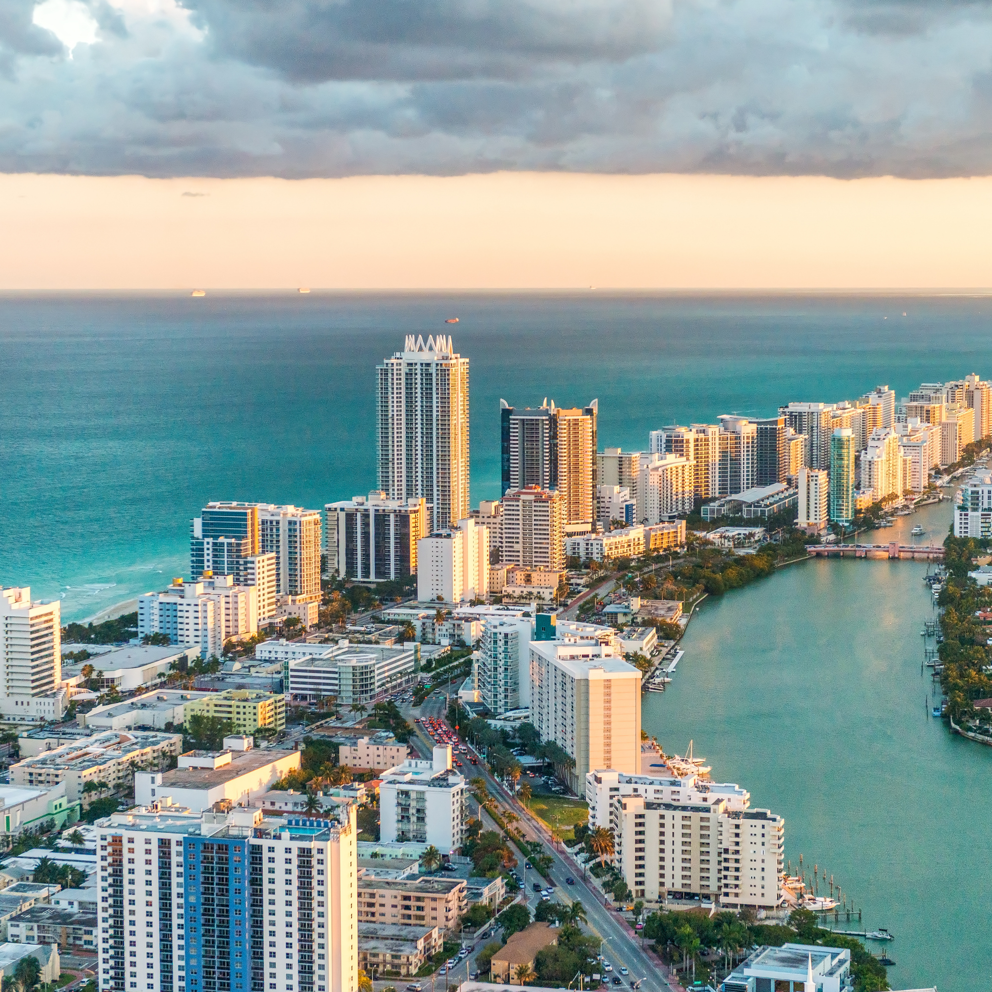 A cityscape with high-rise buildings along a waterfront, showing a mix of modern architecture under a cloudy sky at sunset or sunrise.