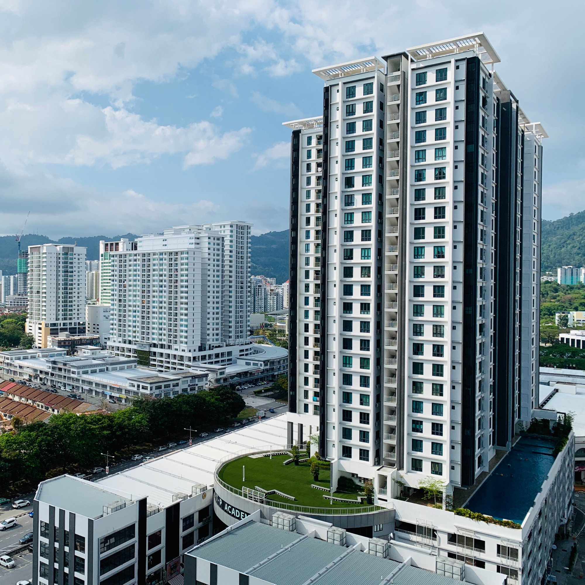 High-rise apartment buildings with a blue sky and scattered clouds in the background, some buildings with white and black facades, green hills in the distance, a parking lot below, and a rooftop garden with trees and grass.