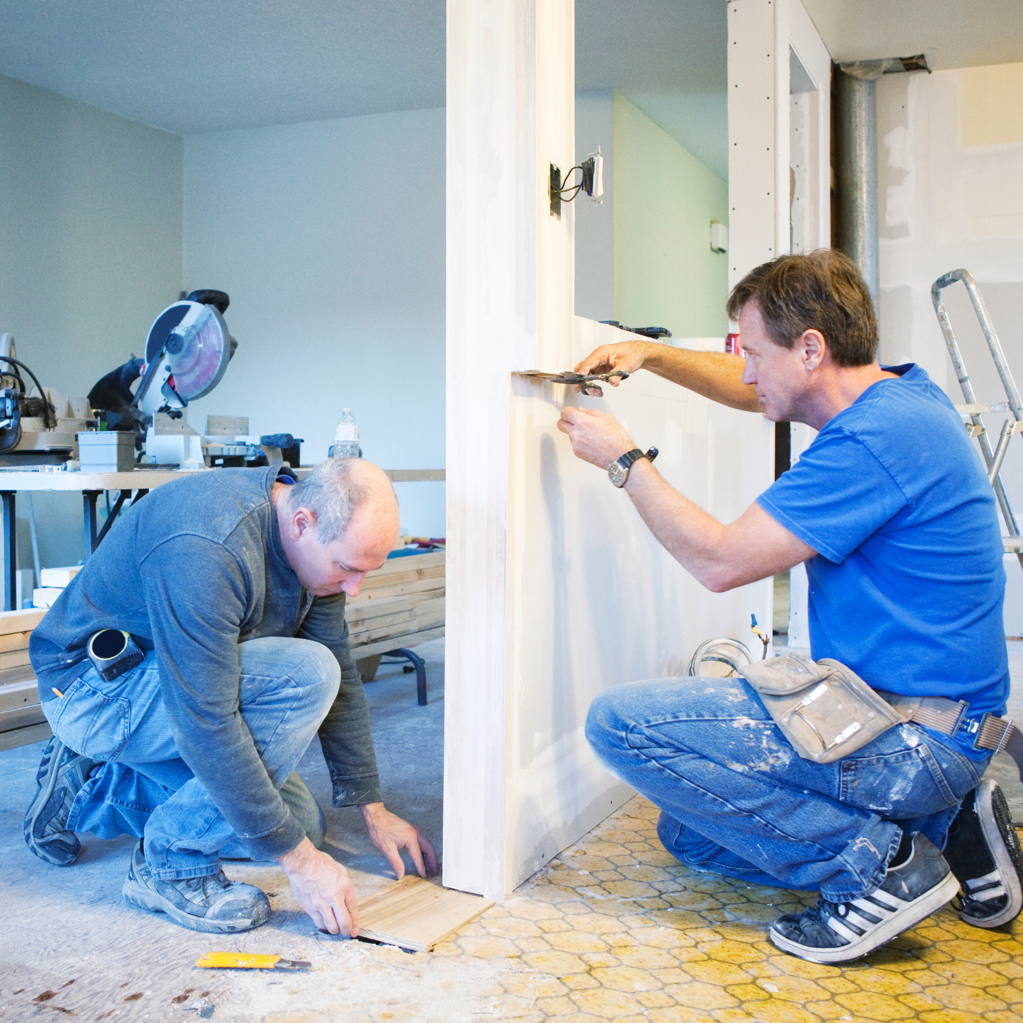 An unfinished room in a house under renovation with supplies and tools, including paint buckets, cardboard, a stepladder, and scattered painting materials on the floor.