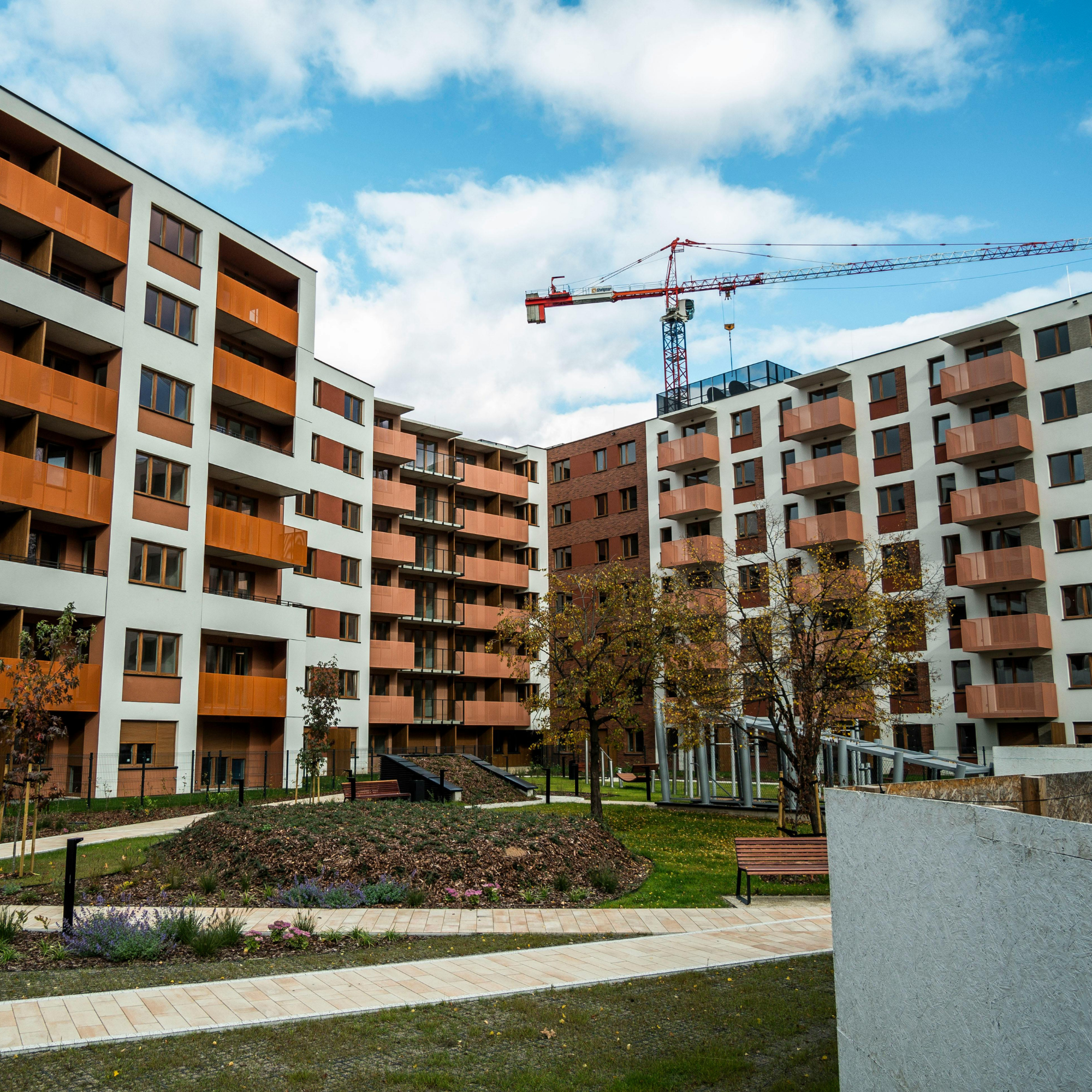 Modern multi-story residential building with balconies, windows, and a rooftop terrace, surrounded by small trees, a pathway, and a grassy area, under a partly cloudy sky.