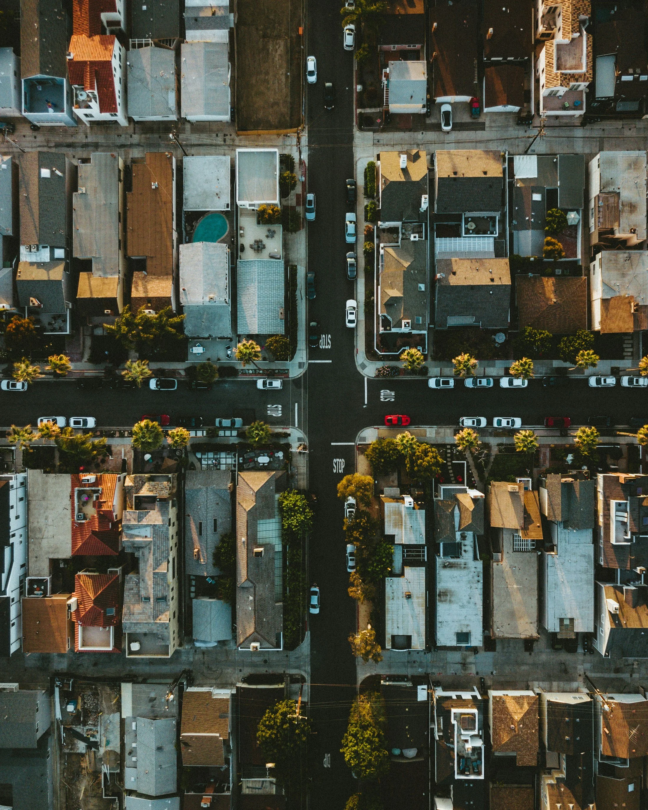 An aerial view of a city intersection with cars, streets, and surrounding residential buildings with rooftops and trees.