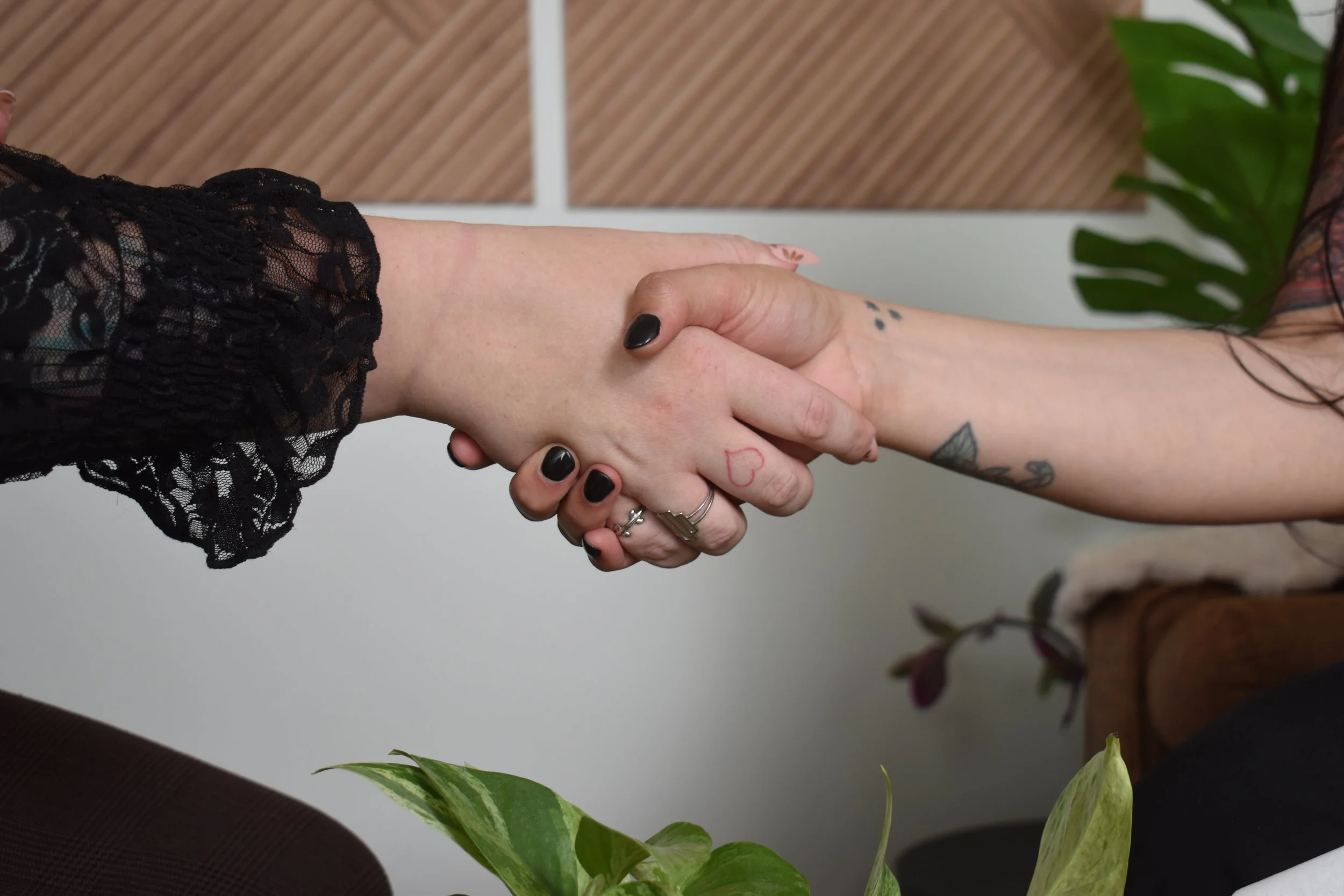Two people shaking hands, one person with black-painted nails and tattoos on the arm, the other with tattooed fingers and a lace black sleeve.