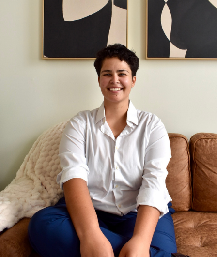 A woman sitting on a brown sofa with a cream-colored blanket, smiling in a room with abstract black and white artwork on the wall behind her.