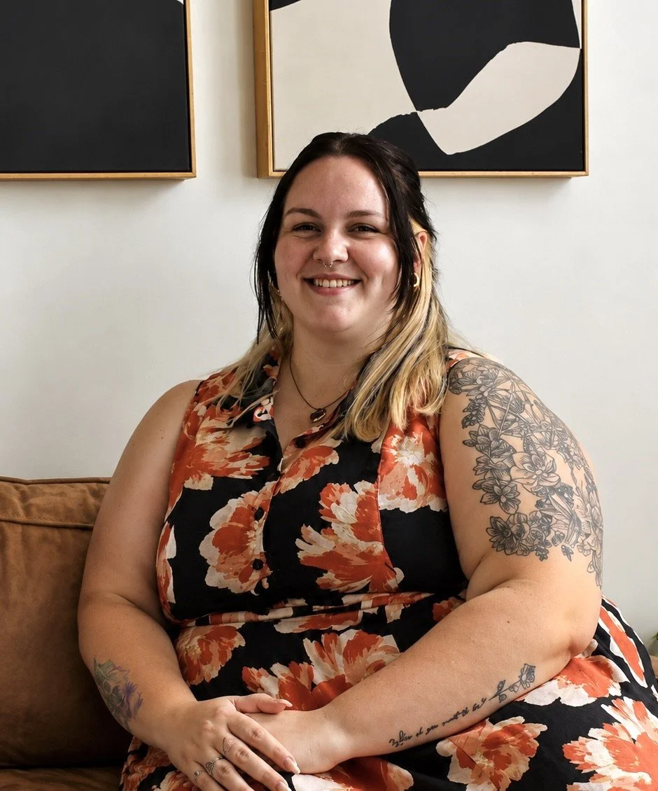 Smiling woman with tattoos, dark hair with blonde tips, seated on a brown couch in front of black and white abstract art.