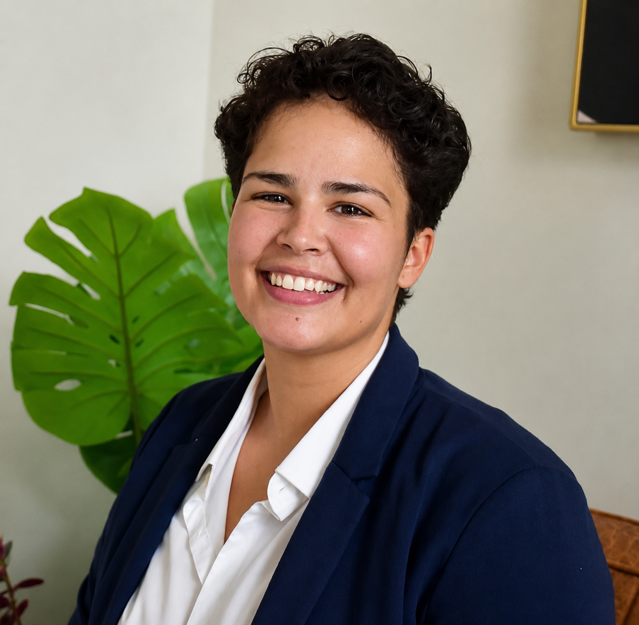 A smiling young woman with short curly dark hair wearing a navy blazer and white shirt posing indoors with a large green leafy plant in the background.