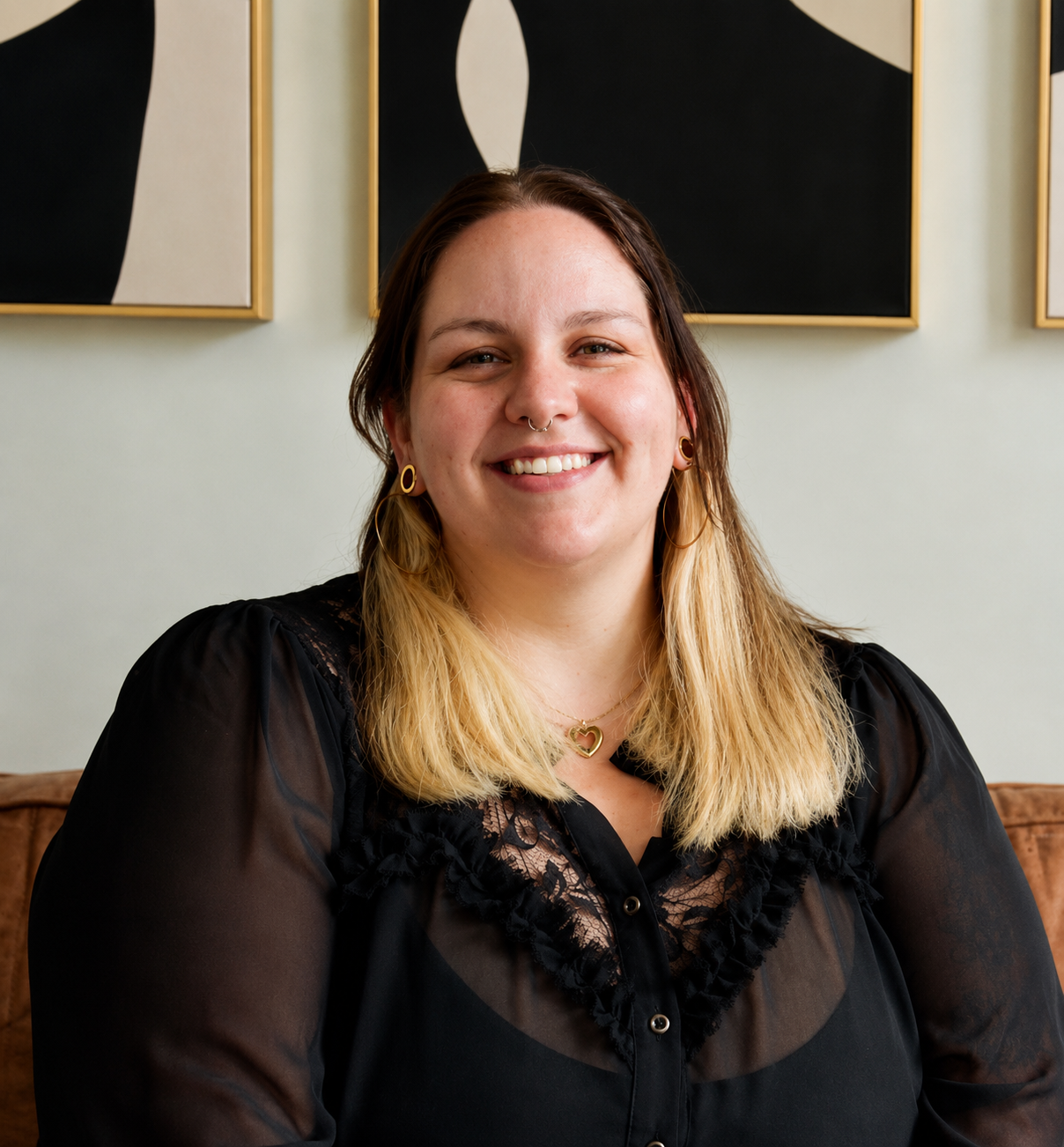 A woman with light skin, shoulder-length blonde hair, and a septum piercing, smiling at the camera, sitting indoors with abstract black and white art on the wall behind her.