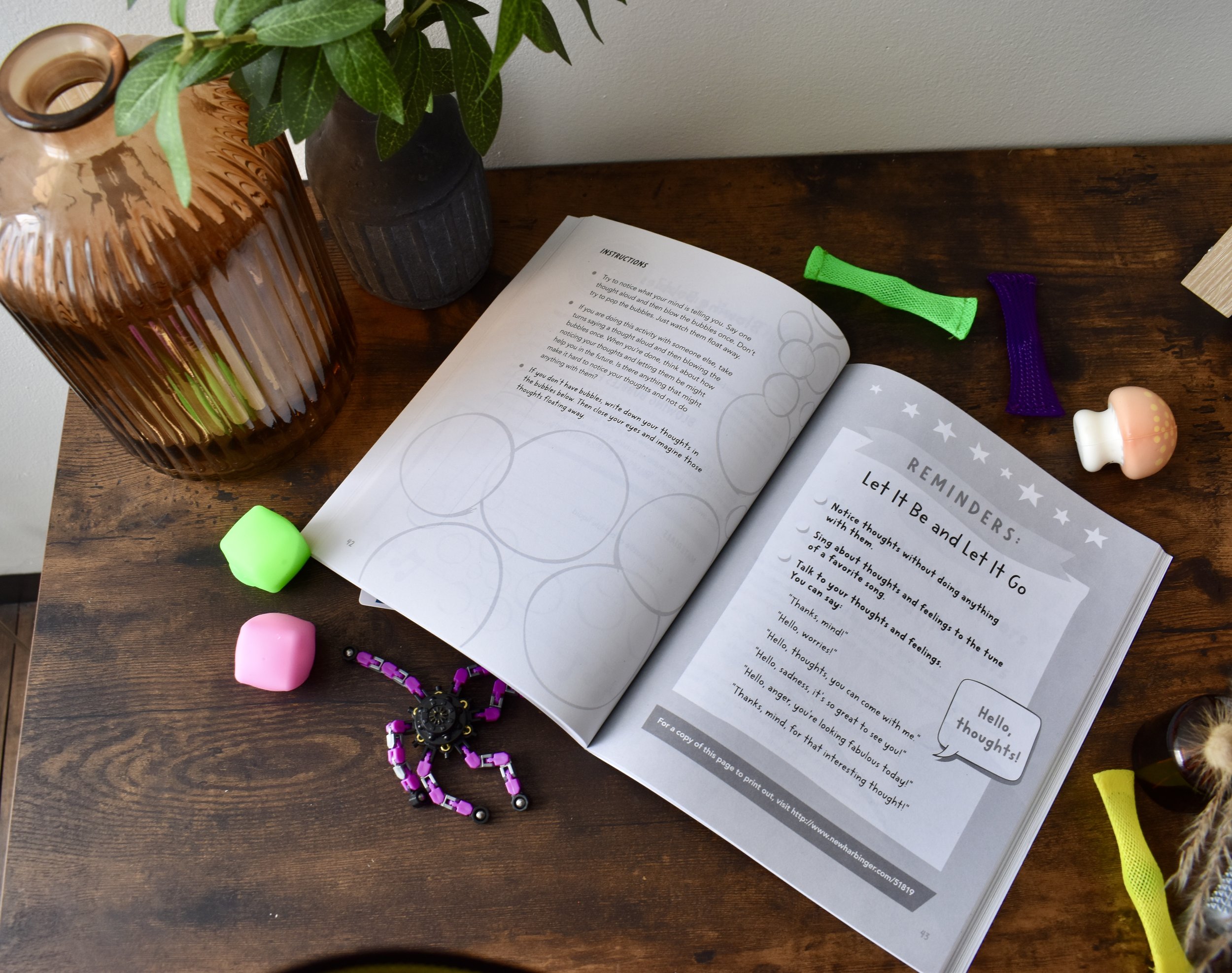 An open booklet with instructions on a wooden table surrounded by colorful items including a ceramic vase with green leaves, a ceramic mug, three foam curlers in green, purple, and yellow, a pink spinning top, a black and purple spider-shaped toy, and part of a cup with a furry animal attached.