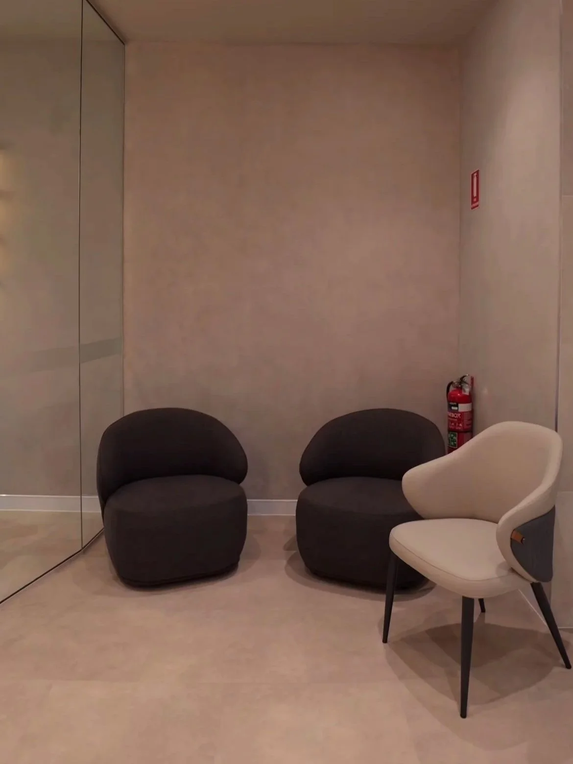 Two black chairs and one white chair in a waiting area with a fire extinguisher near the wall and a mirrored closet door on the left.