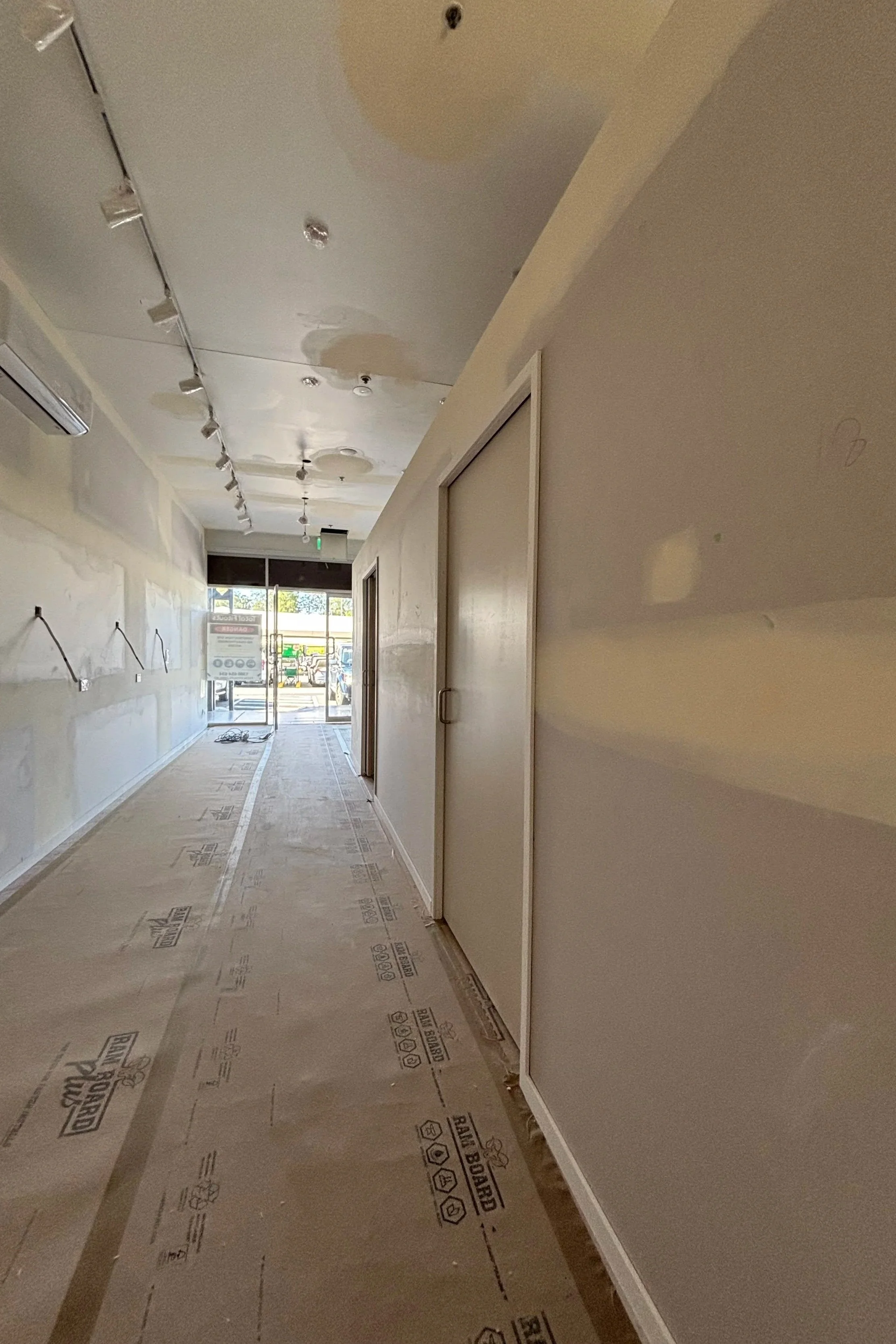 The image shows an indoor corridor likely under renovation, with drywall on the walls and ceiling, some electrical wiring and fixtures, and floor covering still in place. There is a glass door at the end of the corridor, allowing natural light to enter.