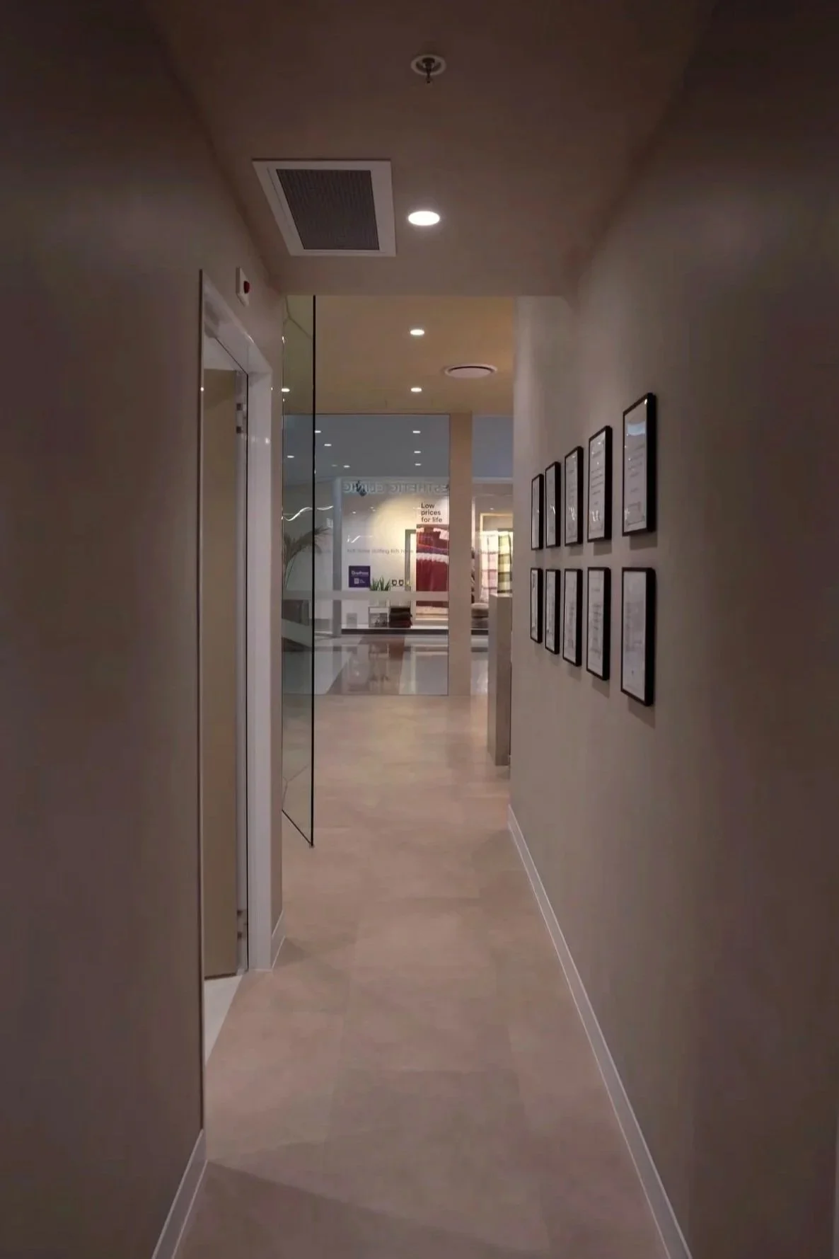 A hallway in a modern building with framed certificates or documents on the right wall. The hallway leads to an area with visible glass doors and signs, including a sign that reads "Low prices for life." The ceiling has recessed lighting and an air vent, and the floor is light-colored.