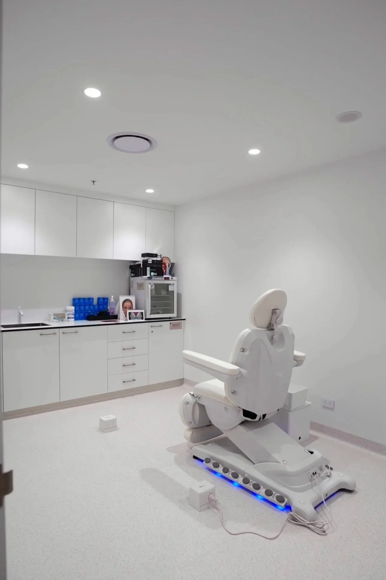 A white medical examination room with a patient chair and medical supplies, including a cabinet with medical items and a framed photo, and a robotic dental or medical chair with a control panel and blue lighting on the base.