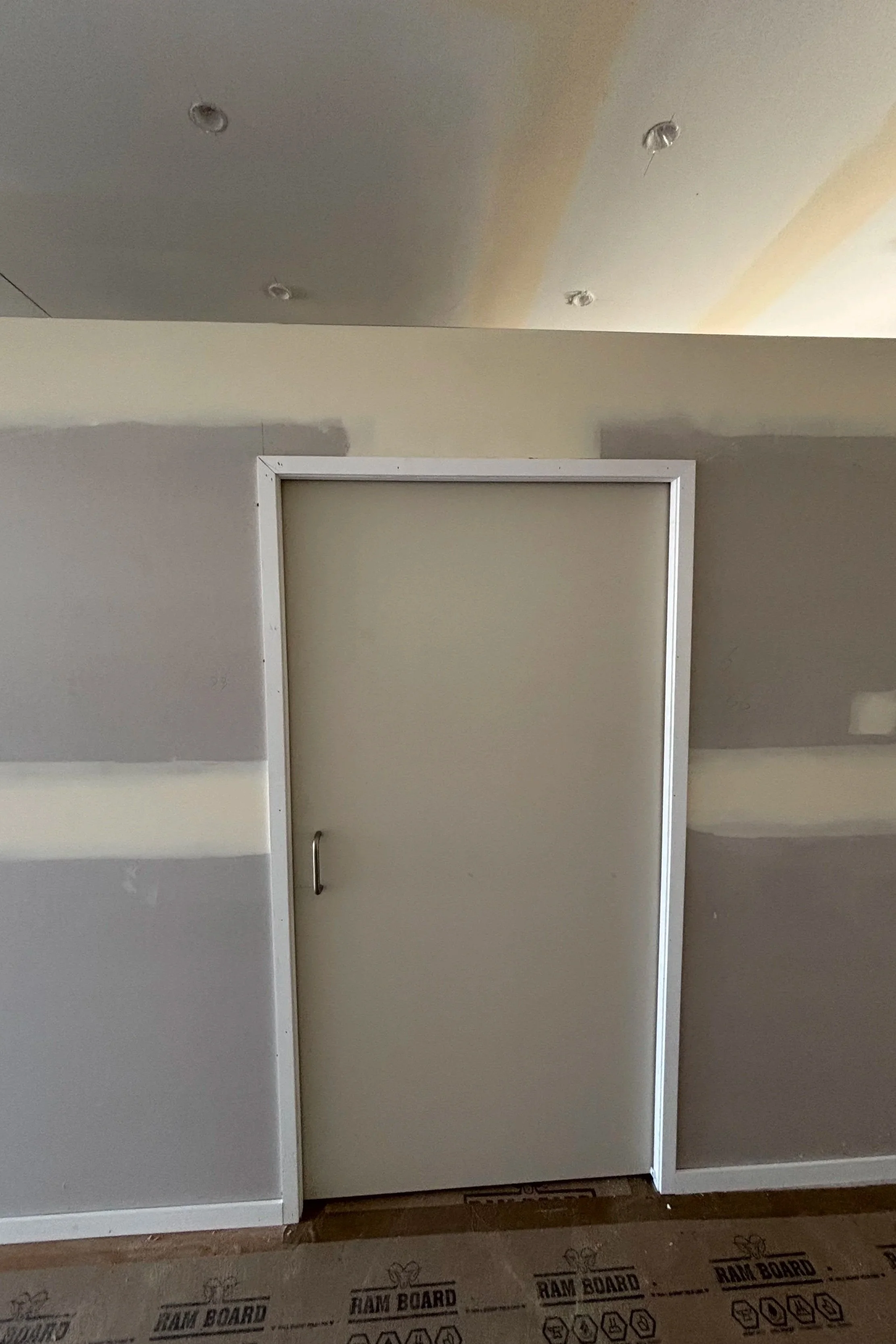 Half-constructed white cabinet with a door, situated against a beige wall with paint patches, and a brown protective floor covering with 'RAM BOARD' printed on it.