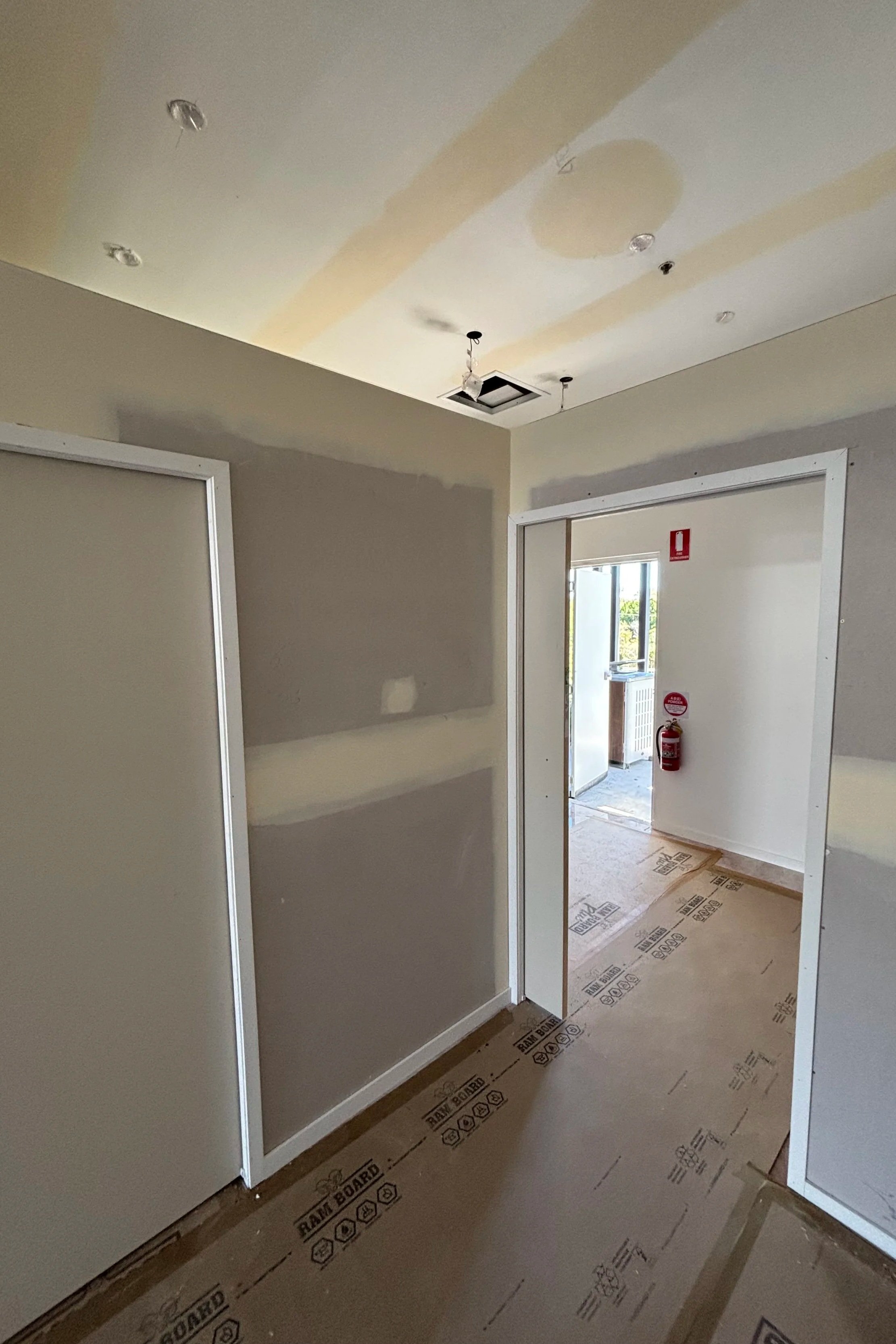 Interior of a room under construction with drywall installed, door frames, protective floor covering, and an open ceiling vent with electrical wiring. Fire extinguisher visible near doorway.