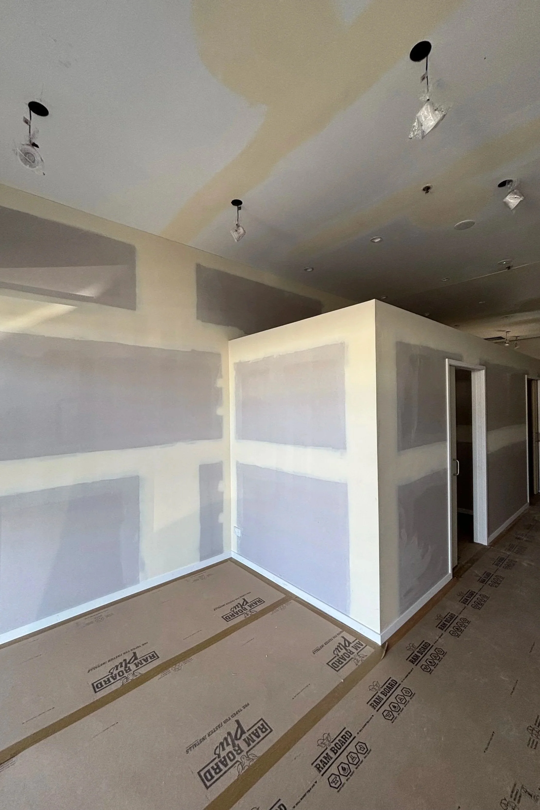 Room with drywall partially painted, ceiling with hanging light fixtures, and flooring covered with protective paper.