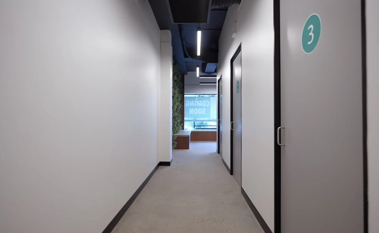 Empty hallway with white walls, black trim, and closed doors numbered 3, leading to a window with a view outside, in a modern building.