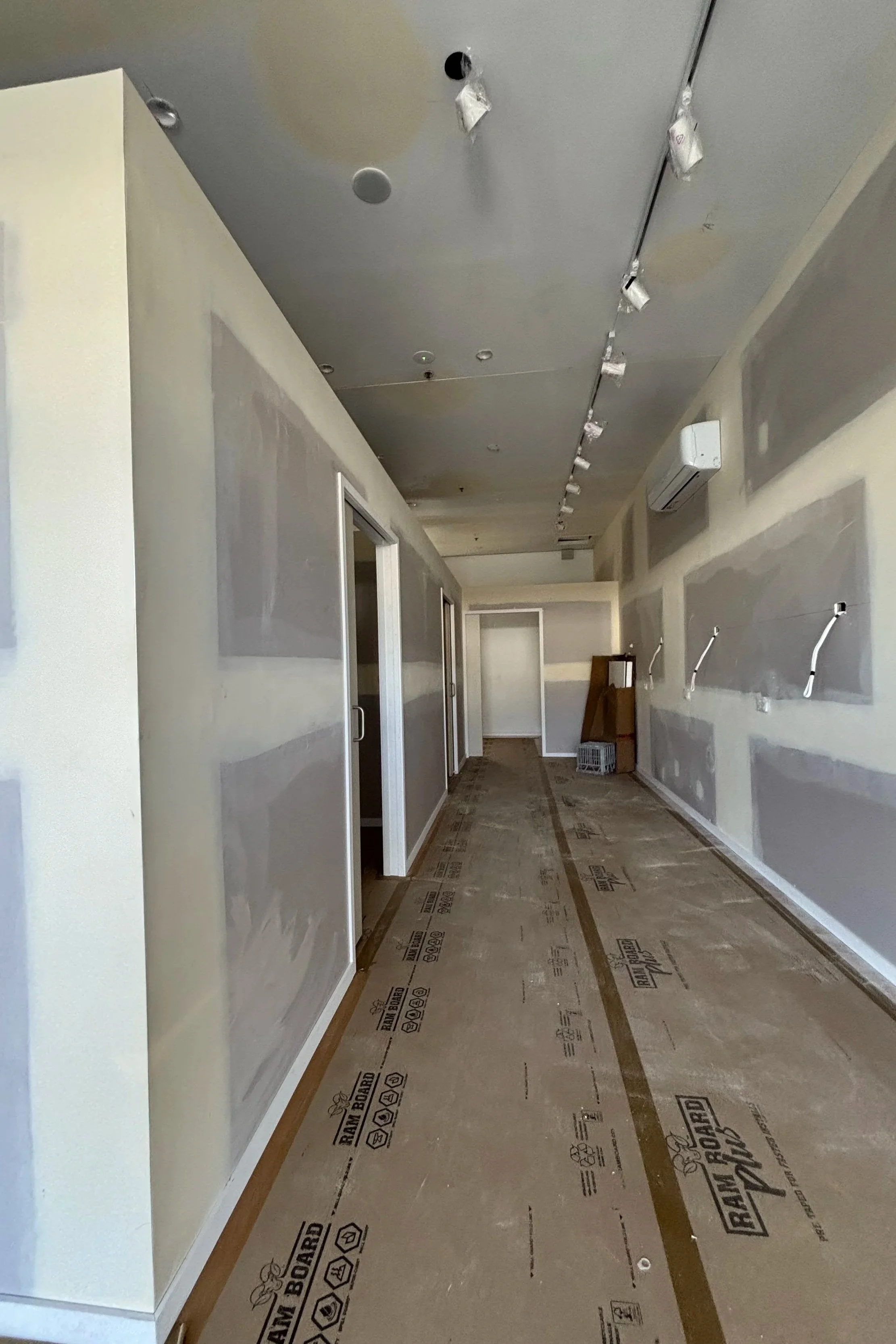 An indoor hallway under construction with a protective floor covering and unfinished walls, featuring a white air conditioning unit and several doorway openings.