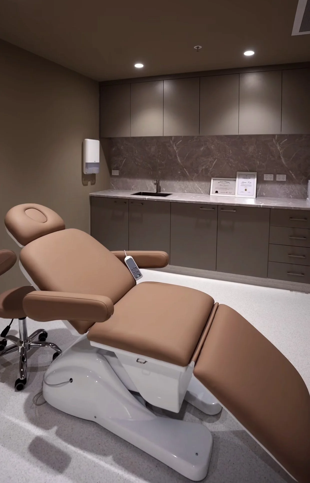 Medical examination room with a brown reclining chair in the foreground and a kitchen-like area with cabinets, a sink, framed certificates, and a wall-mounted sanitizer dispenser in the background.