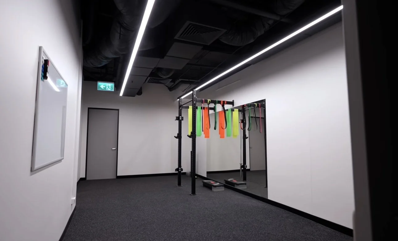 Empty fitness room with black ceiling, white walls, gray carpet, and a black metal frame with colorful resistance bands hanging, and a mirror on the wall.