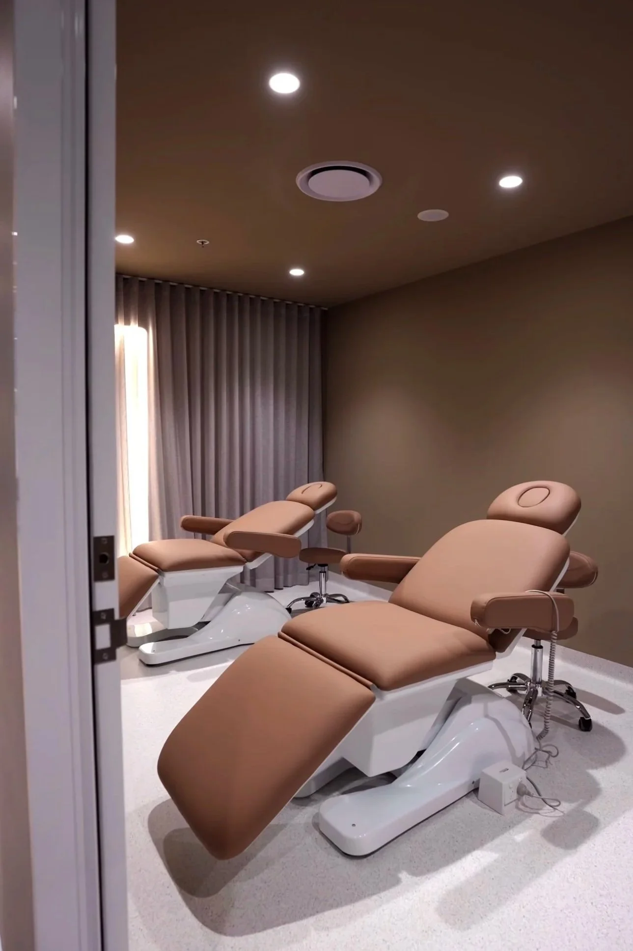 Three beige medical recliner chairs in a room with beige walls, grey curtains, and ceiling recessed lighting.