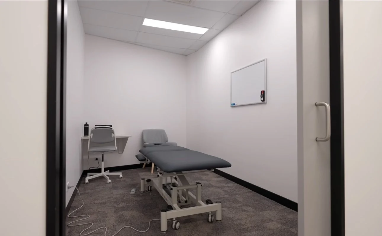 Empty medical examination room with a bed, white walls, a whiteboard, and a small desk with a chair.
