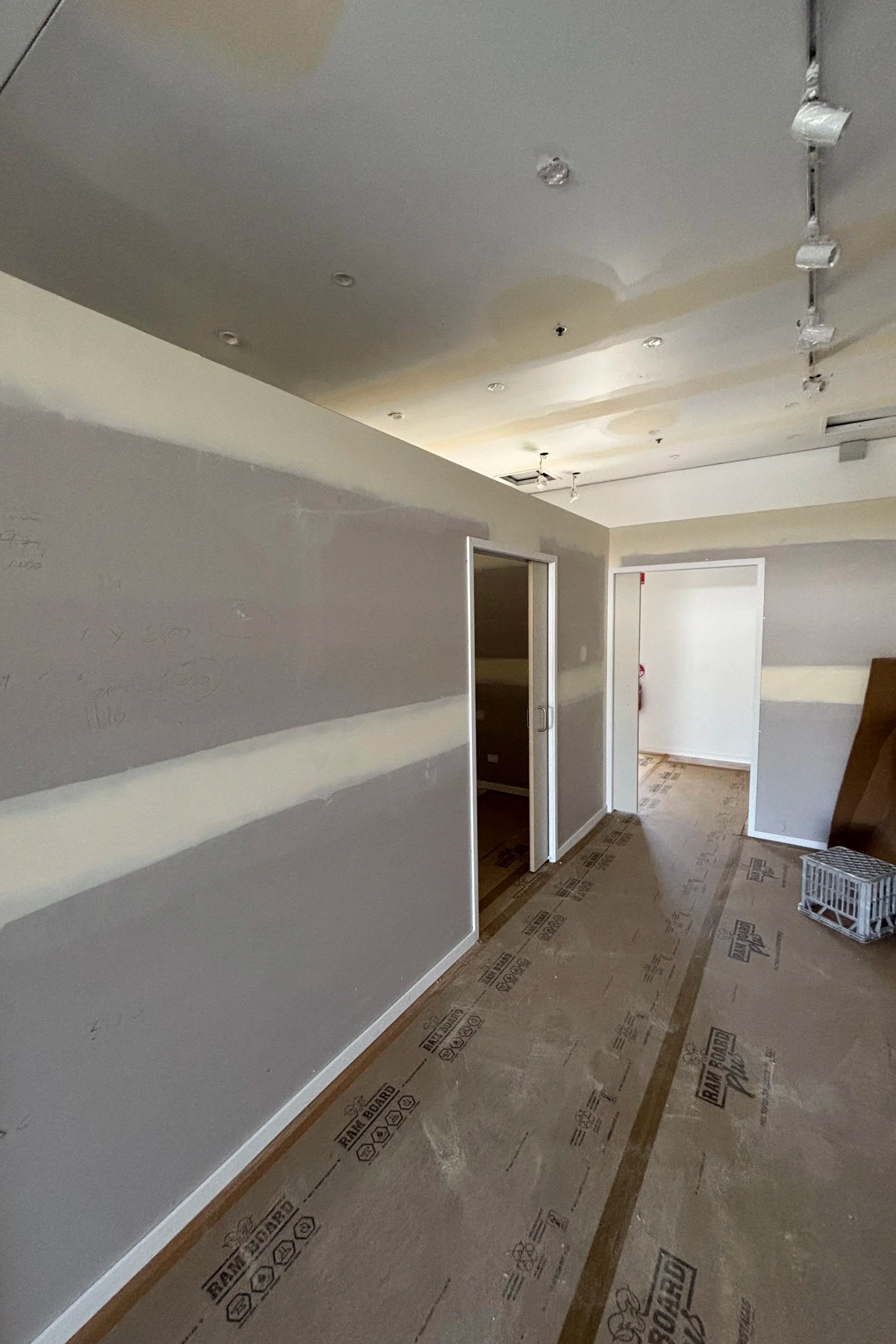 Interior of a room under construction with unfinished flooring, partially painted walls, ceiling lights, and doorways.