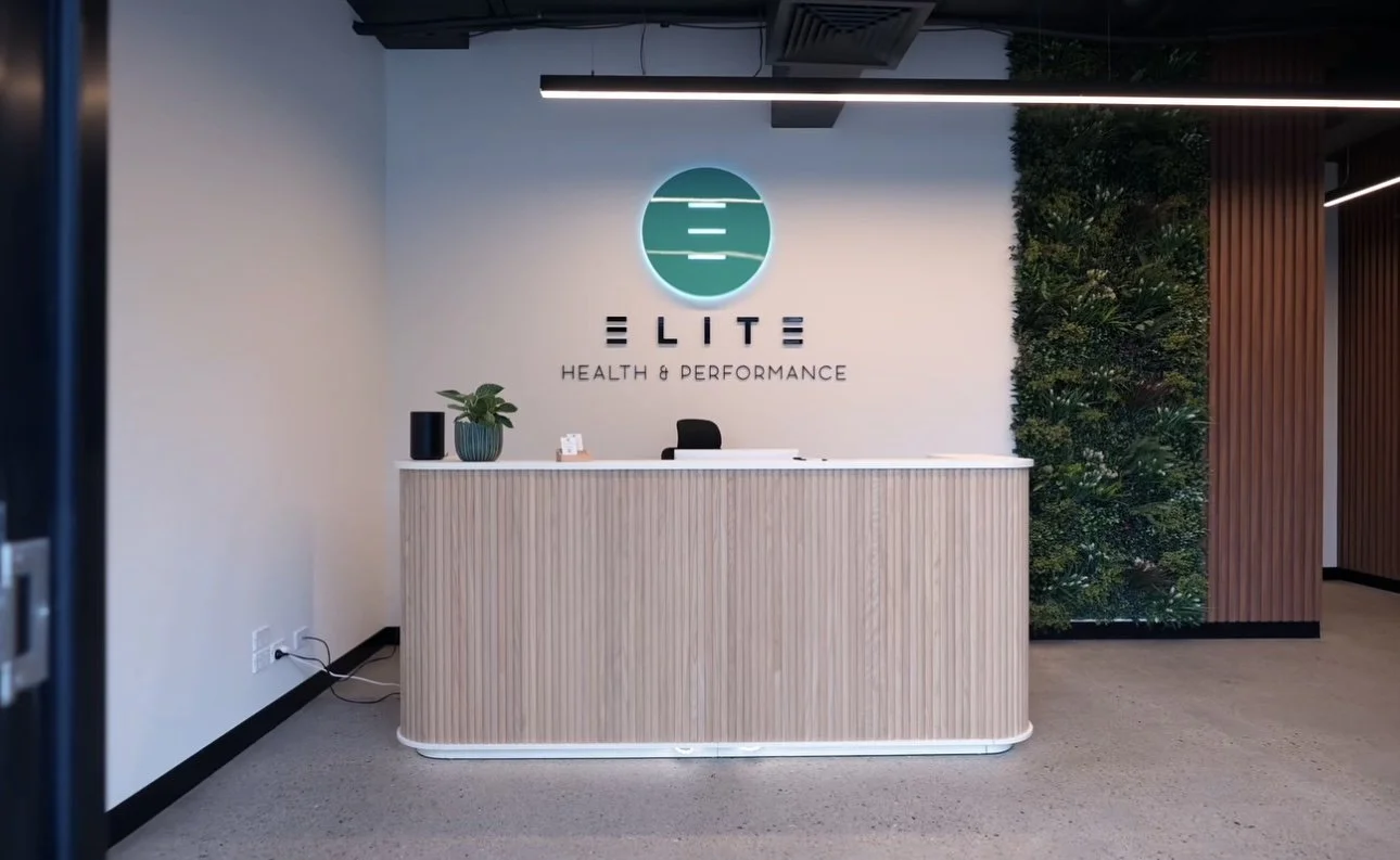Reception desk with a green plant and black speaker on it, behind which is a sign that says "ELITE HEALTH & PERFORMANCE" with a circular logo above, in a modern office lobby with wooden wall accents and a green wall of plants.