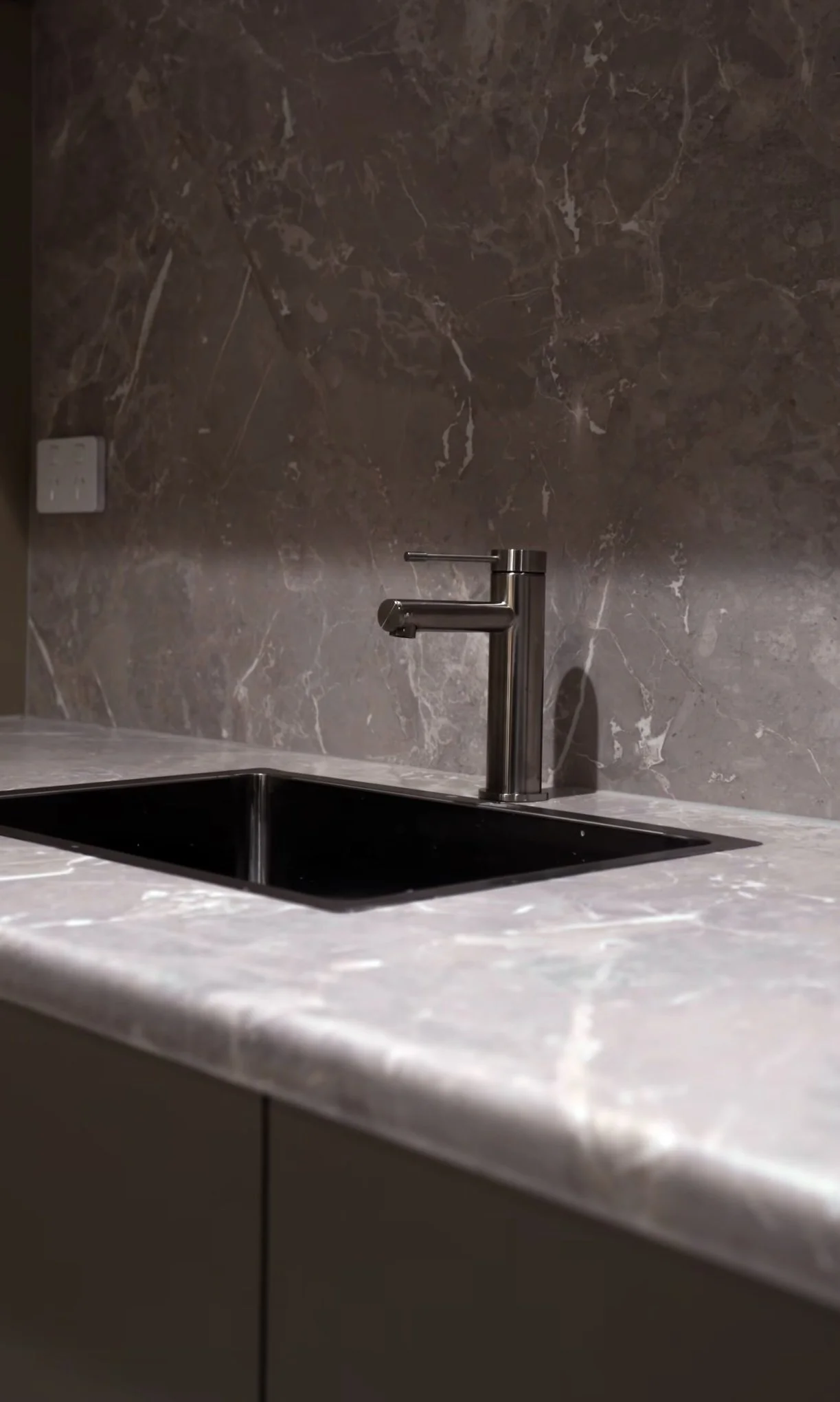 A modern kitchen sink with a matte stainless steel faucet, set against a dark marble backsplash and a white marble countertop.