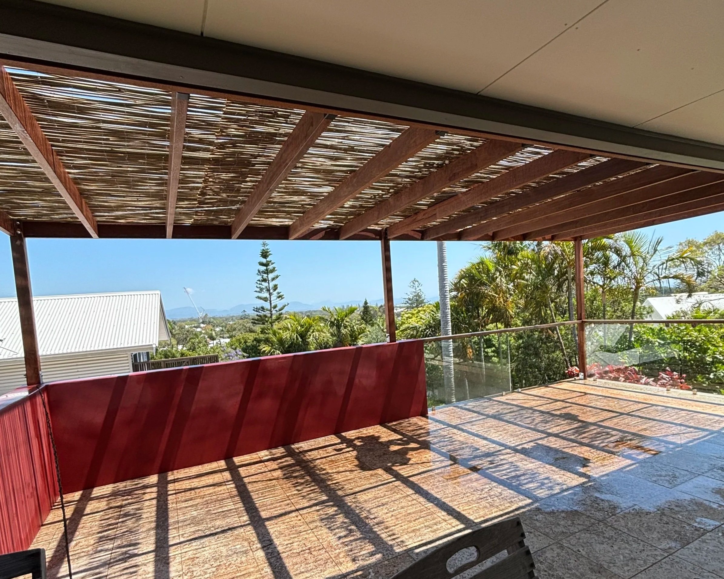 View from a balcony or patio with a wooden pergola overhead casting striped shadows, overlooking a garden with palm trees, bushes, and a building with a metal roof, under a clear blue sky.